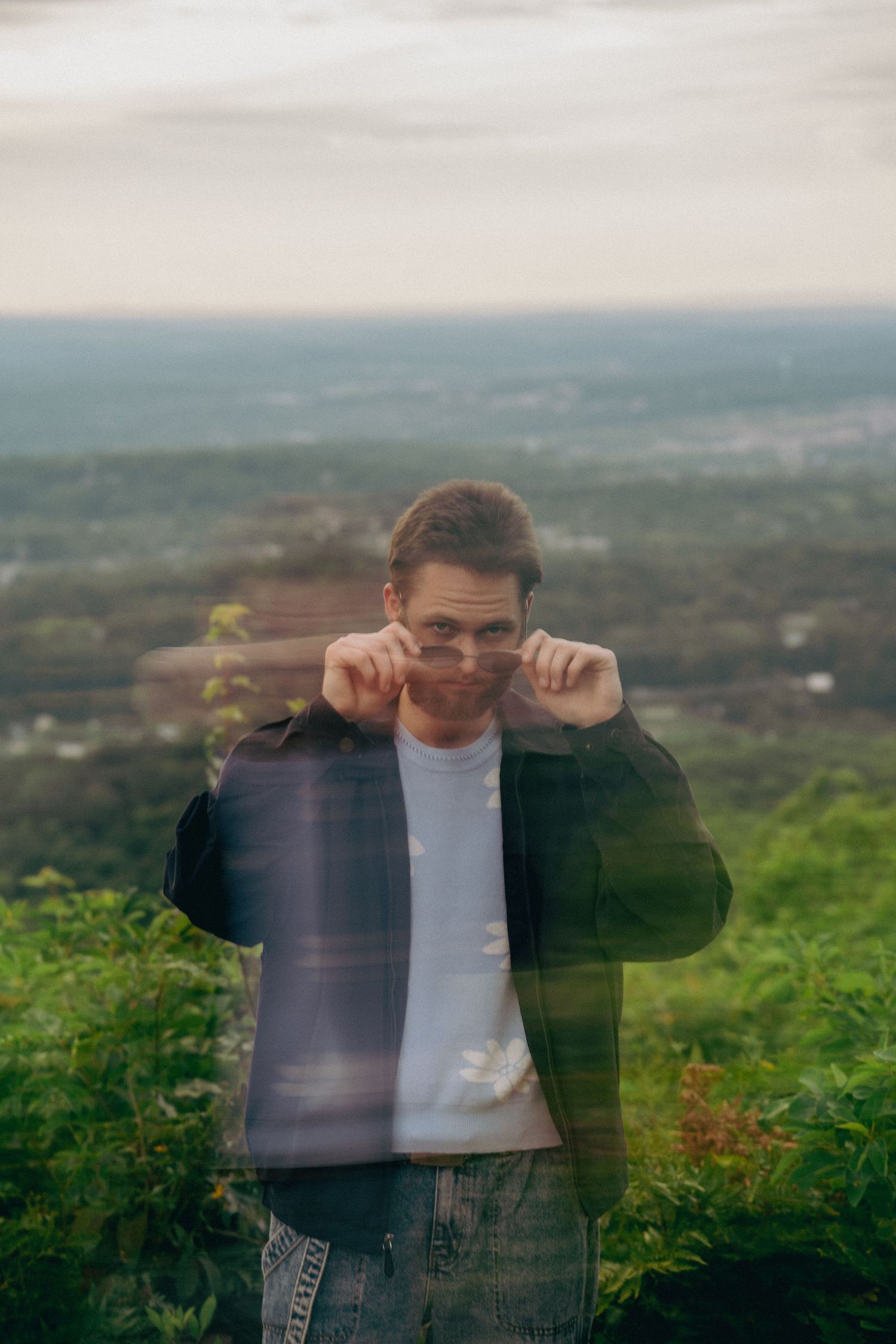 Motion blur captures figure in black jacket against scenic hilltop vista with green foliage in foreground.