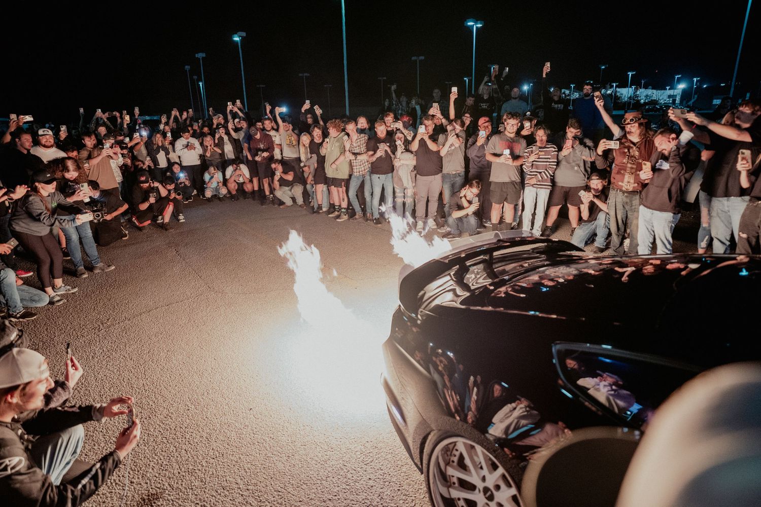 A crowd of spectators gathered around a car at night with the car shooting flames from the exhaust illuminating the scene.