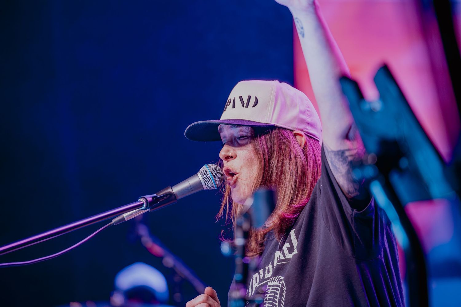 Female vocalist performing live on stage, wearing sunglasses and a cap, singing into a microphone with arm raised.