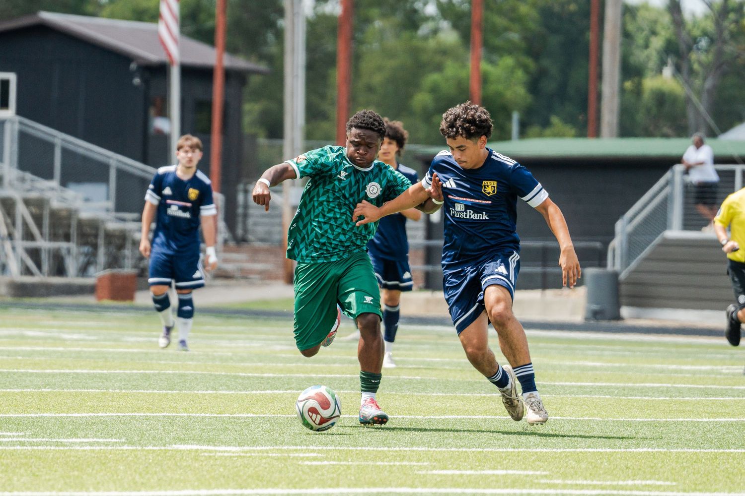 Two soccer players compete for the ball during a match on a green turf field, one in green and one in navy blue.