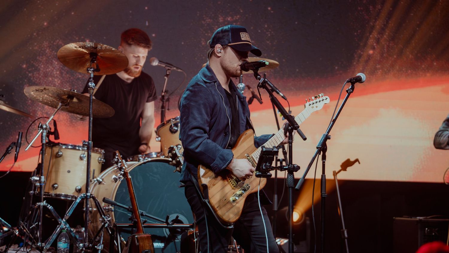 Concert performers on stage with instruments during a live music performance in dramatic red lighting.