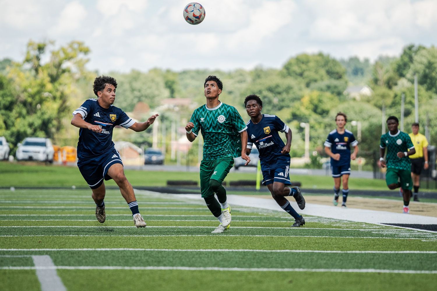 Soccer players compete for the ball during an outdoor match on a green turf field with trees in the background.