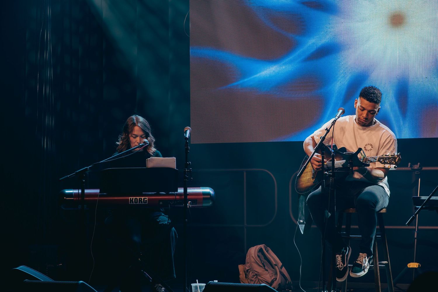 Musicians perform on stage with one playing keyboard and another on acoustic guitar against a blue-lit backdrop.