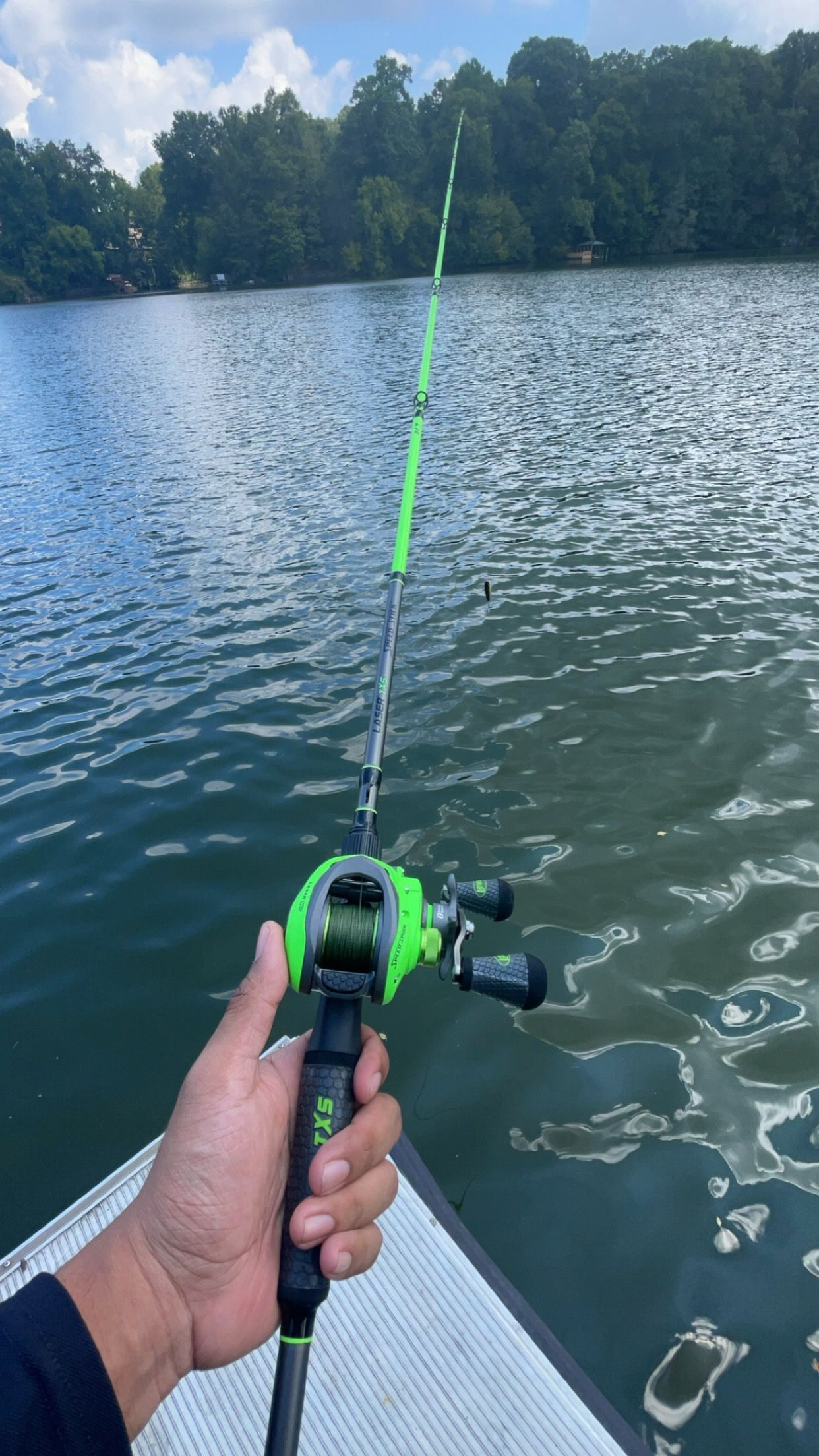 Hand holding a green and black baitcasting fishing rod over calm lake waters with trees in the background.