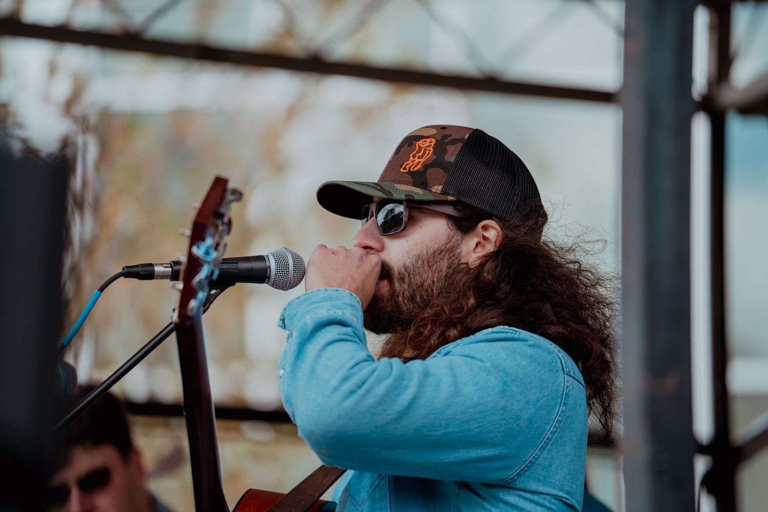 A musician with long curly hair and a beard performs on stage wearing a blue jacket and baseball cap.