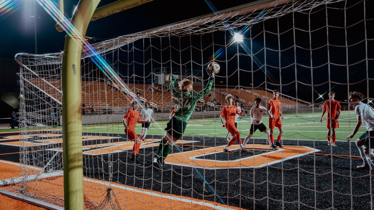 Goalkeeper in green leaps to make a save during a night soccer match, with players in orange jerseys competing near the goal.