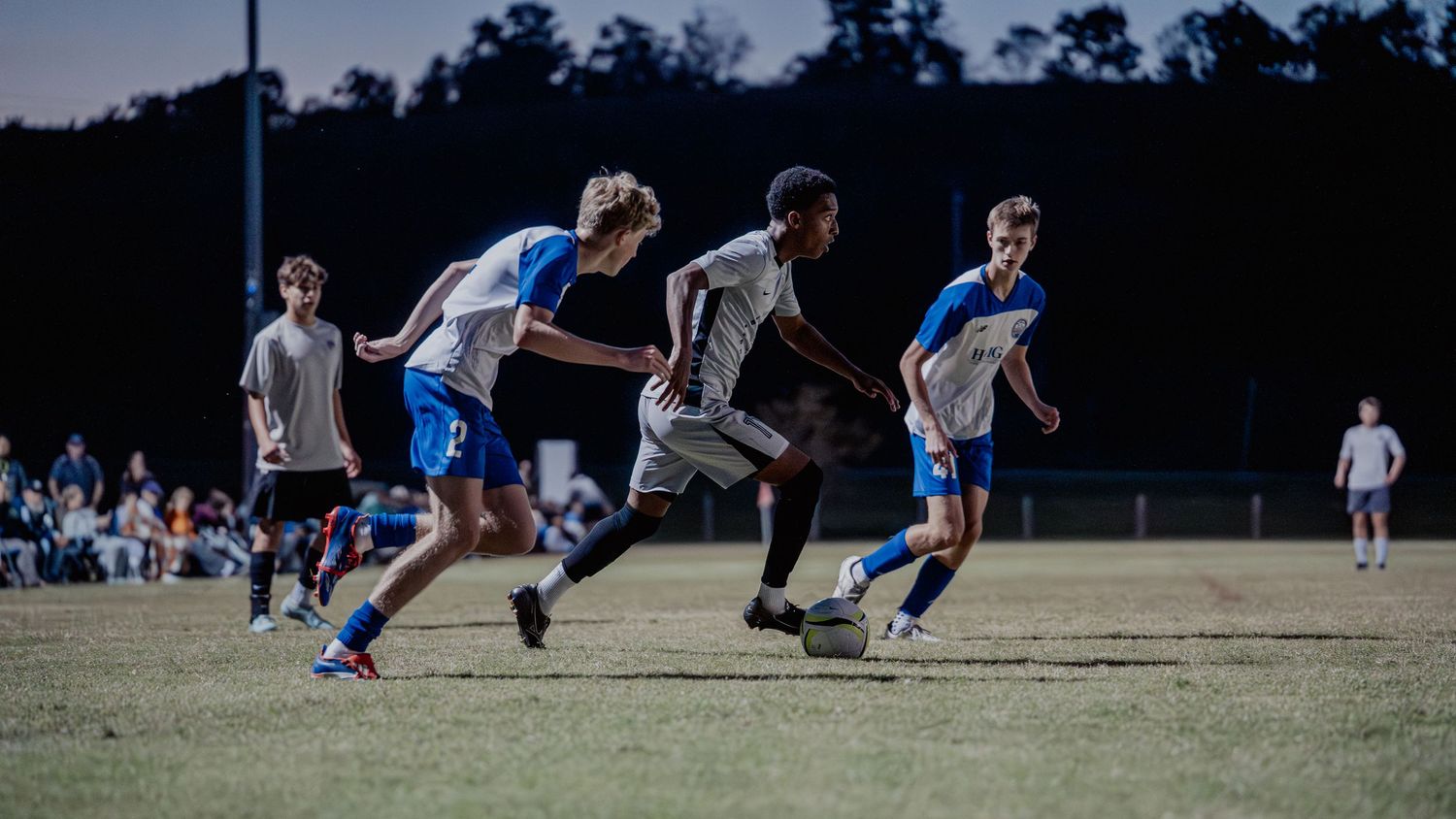 Soccer players compete for the ball during an intense match on a grass field at dusk.