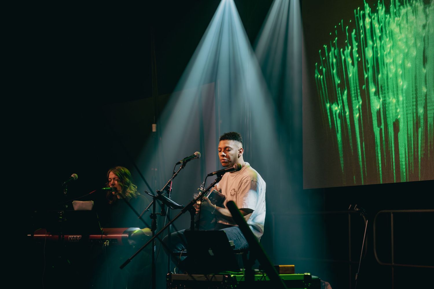 Musician performs on stage with dramatic white spotlights and green projection screen in background during live concert.