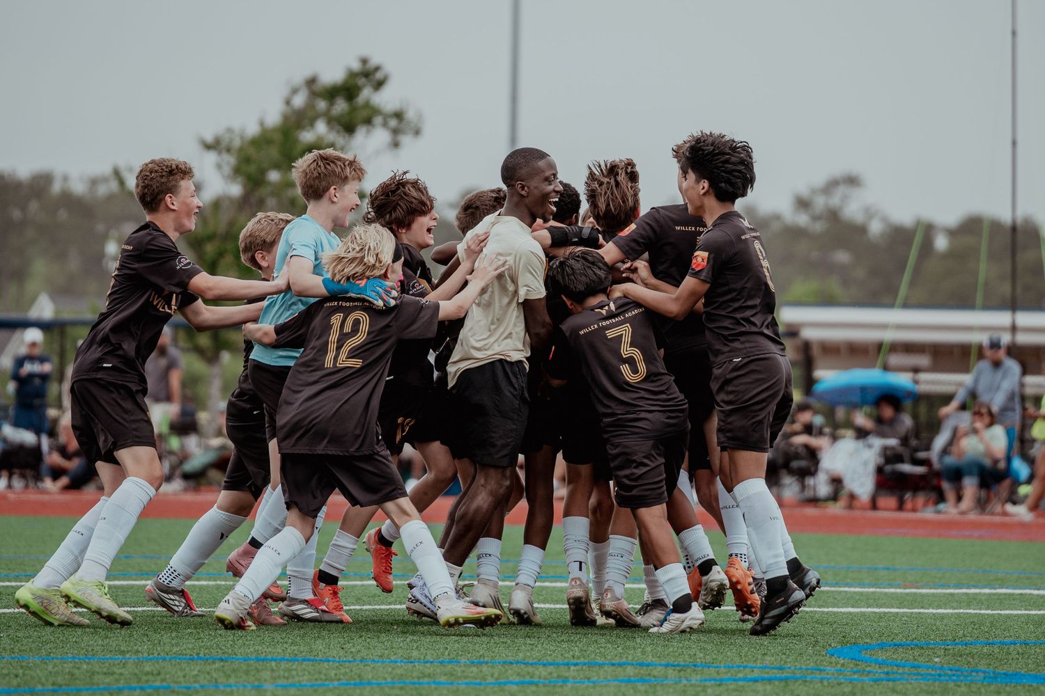 Youth soccer team celebrating a victory on the field, wearing black uniforms, with players huddling together in excitement.
