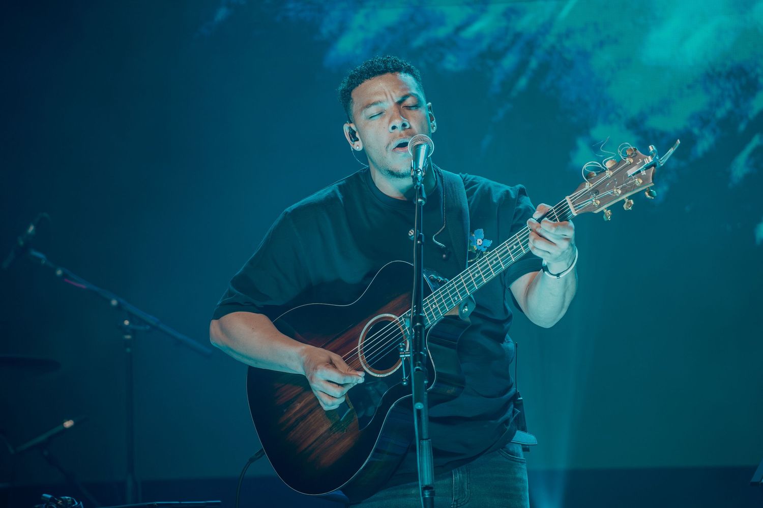 Man in black t-shirt playing acoustic guitar on stage under blue stage lighting.