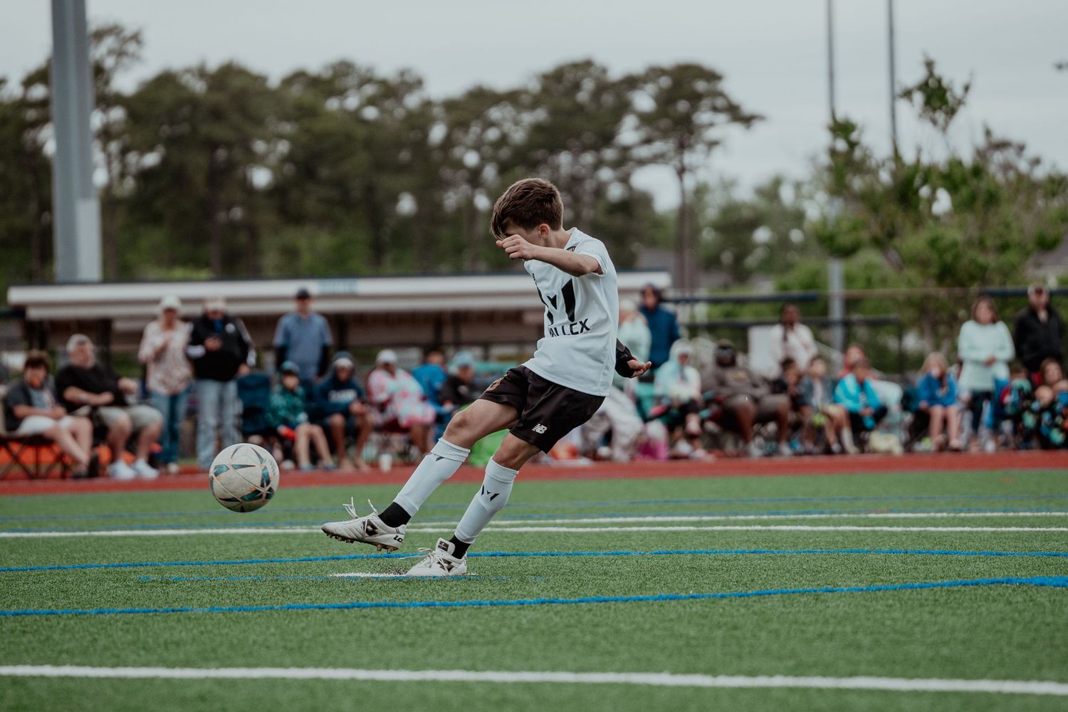 Young boy in white soccer uniform kicking a ball on a turf field during a youth soccer game.
