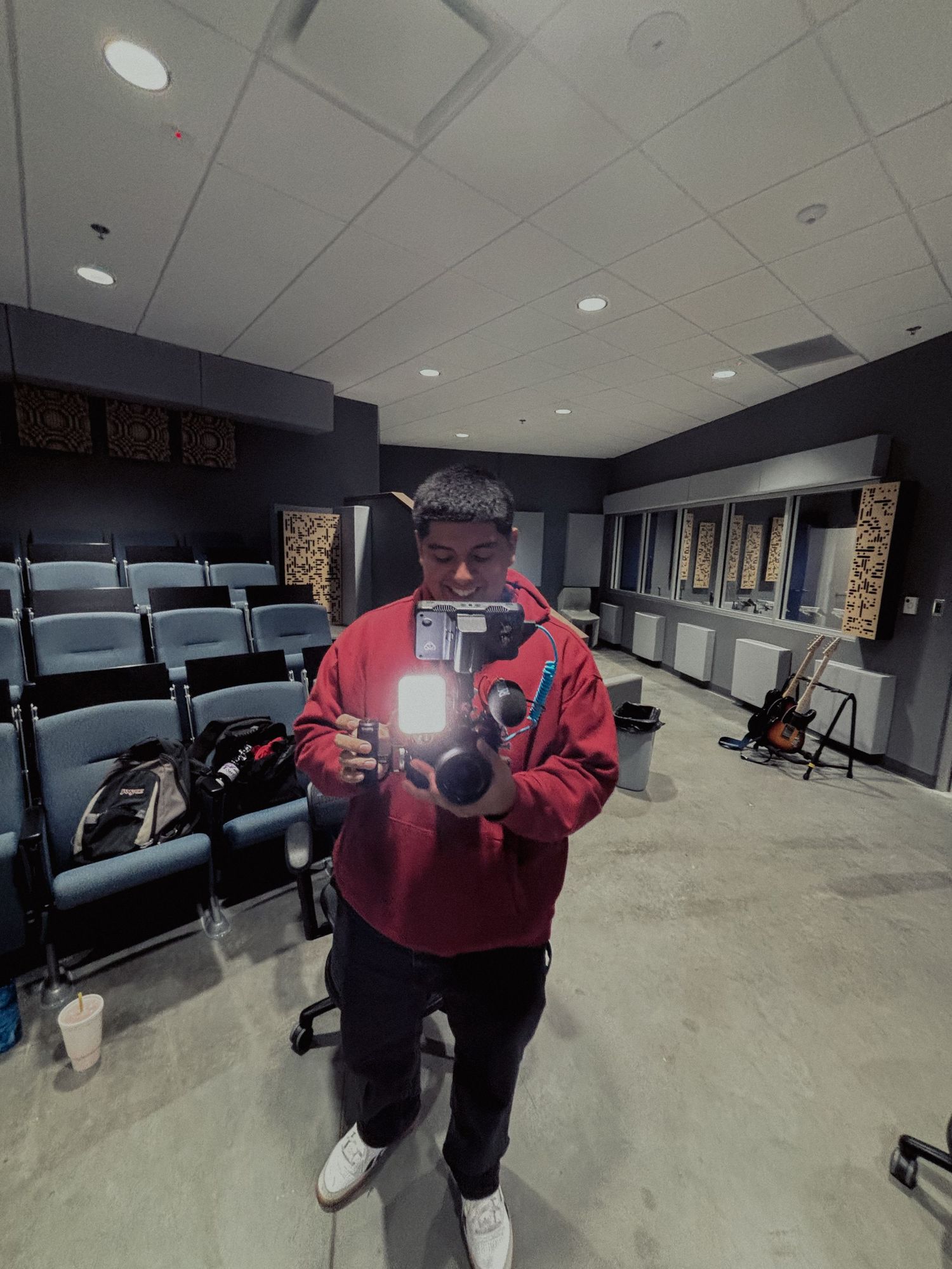 A person in a red jacket taking holding a video camera rig inside a studio classroom.
