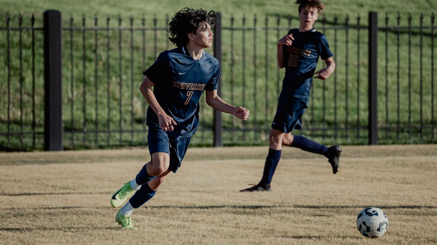 Two youth soccer players in blue jerseys practice on a dirt field with a wrought iron fence in the background.