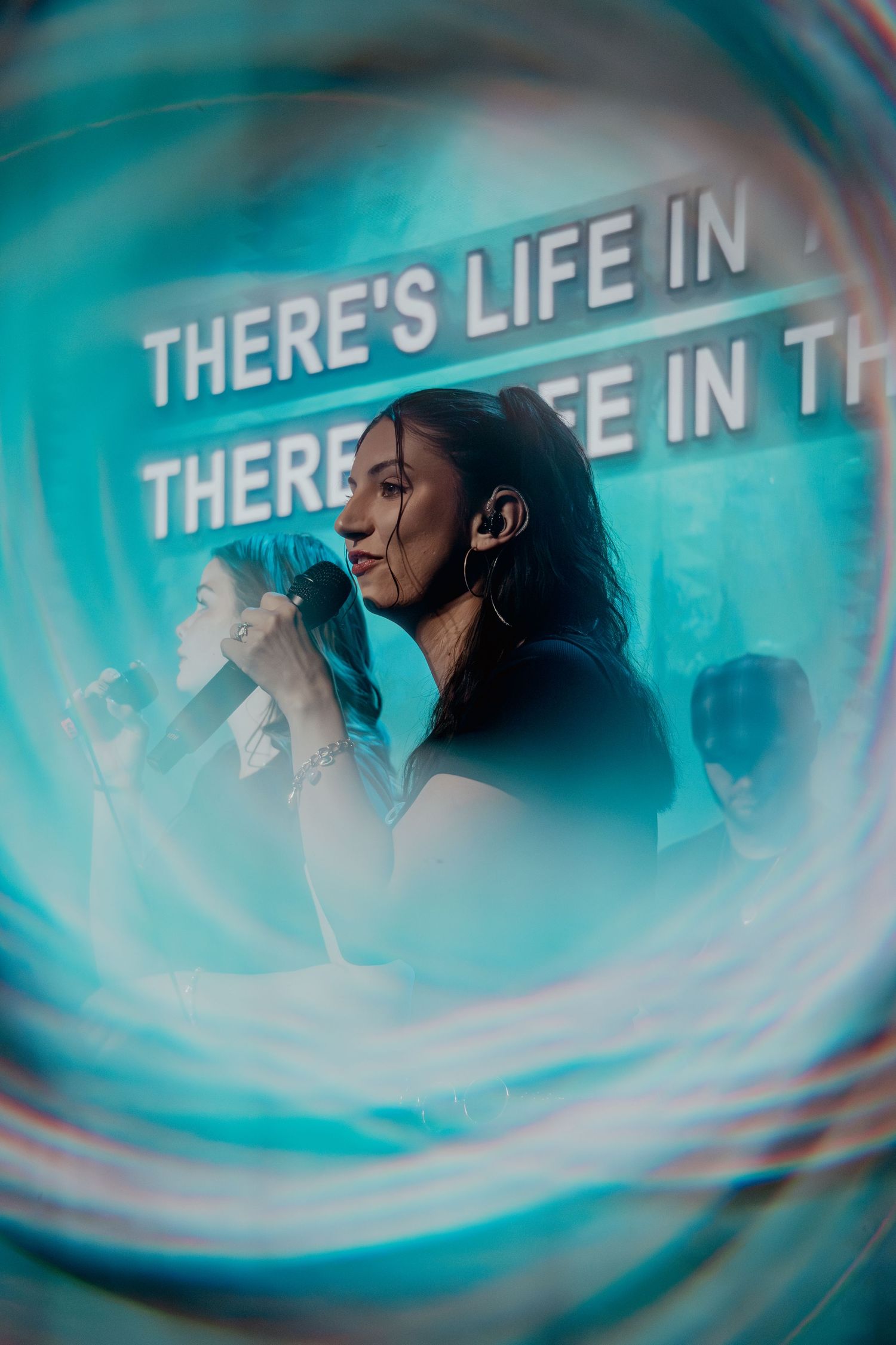 Woman singing into microphone on stage with teal light bokeh effect and 'There's Life In' text backdrop.