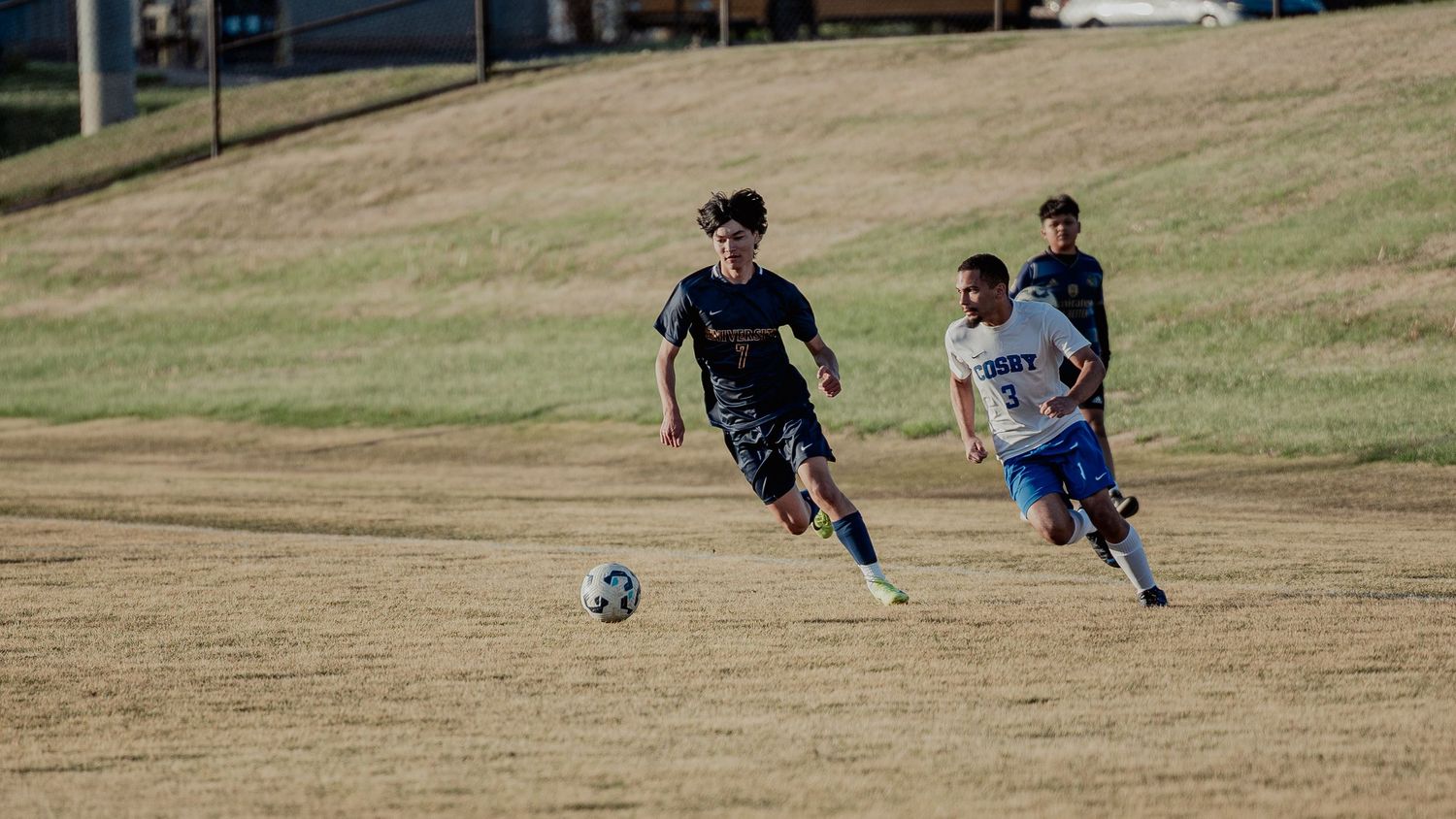 Soccer players chase after the ball during a competitive match on a dry grass field.