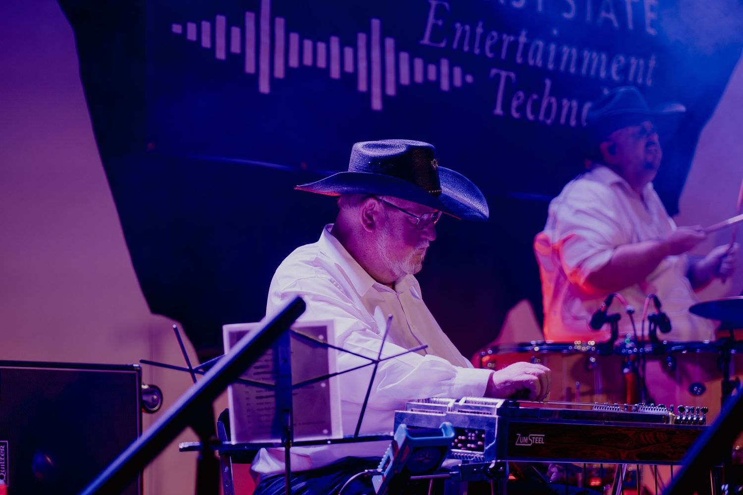 Musician in a cowboy hat performs on steel guitar at a concert with purple stage lighting.