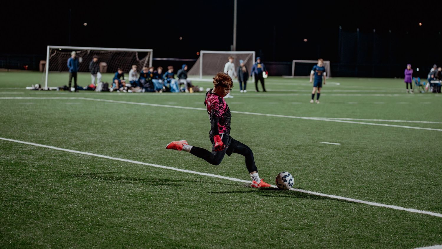 A soccer player in a purple jersey slides to kick the ball on an illuminated turf field at night.