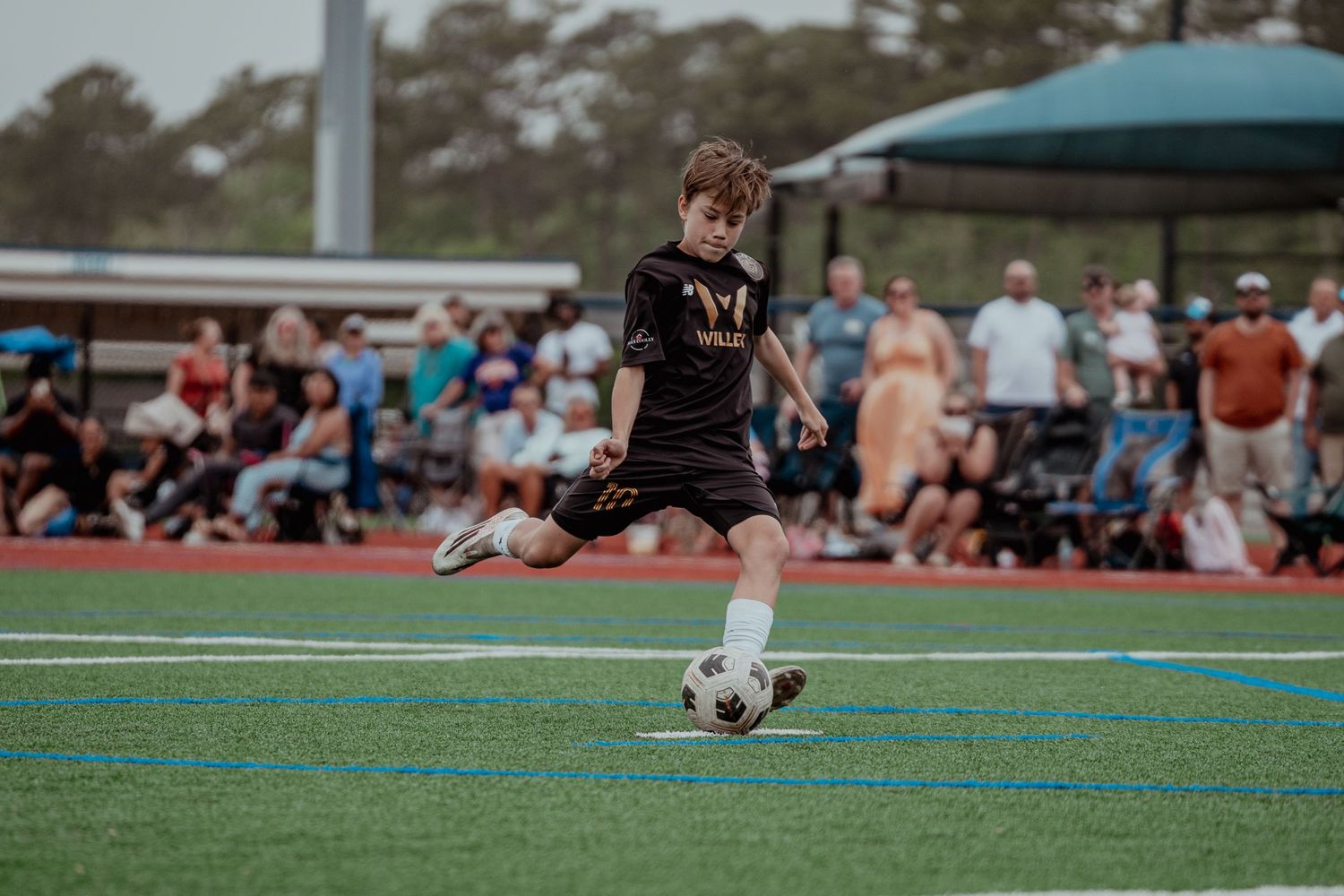 Young soccer player in black uniform kicking ball on blue turf field during competitive match with spectators.