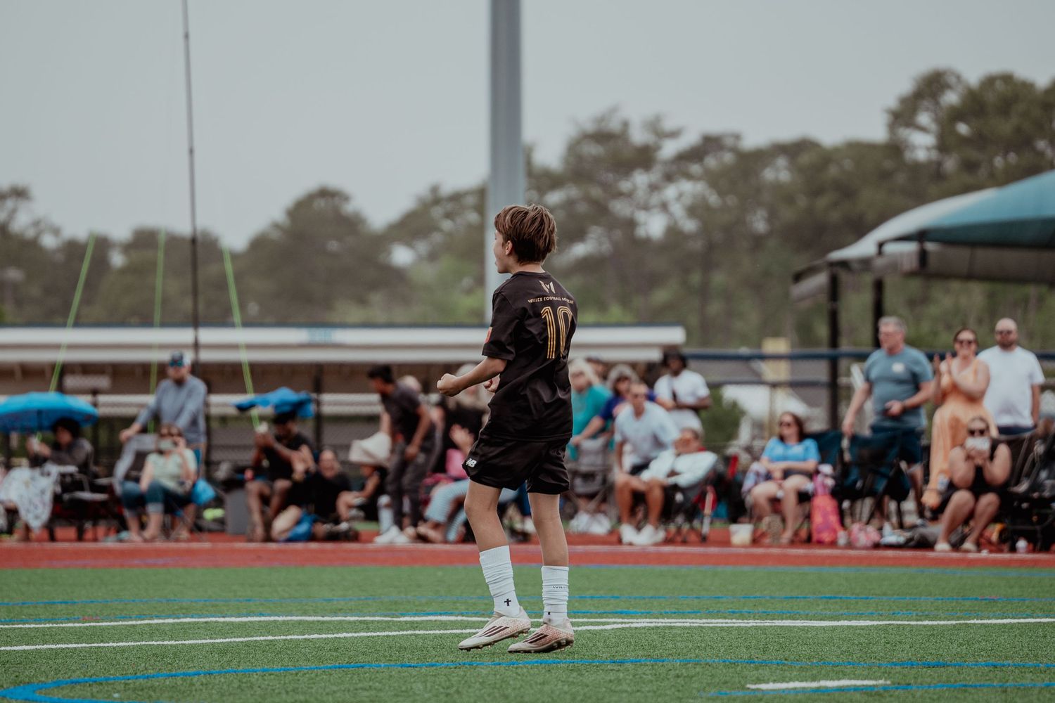 Young soccer player in black uniform stands on red turf field during a game with spectators watching.