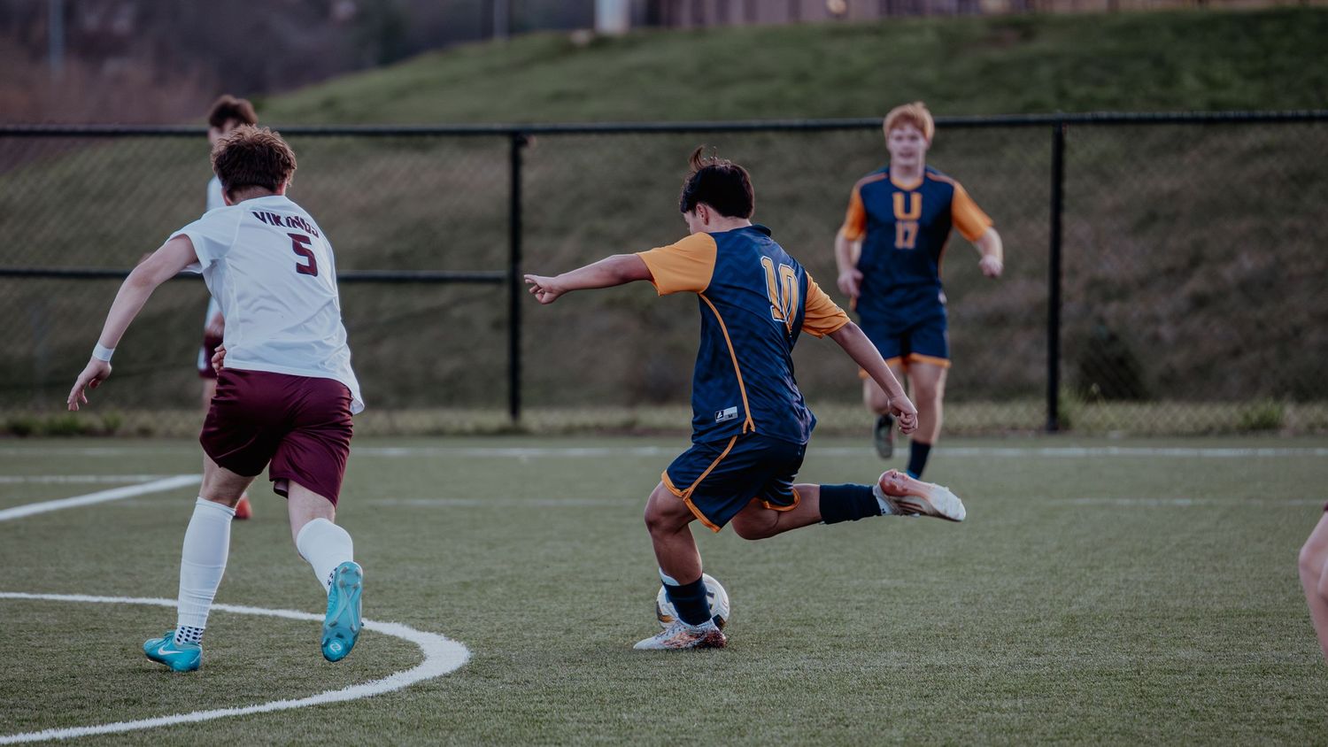 Youth soccer players compete on a green field, with one player in blue and gold kicking the ball.
