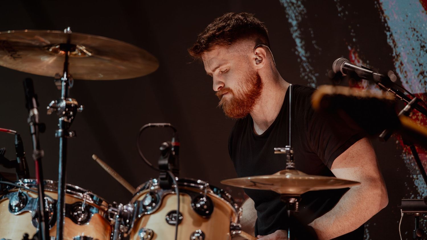 A drummer performs on stage with cymbals and drums while colored stage lights illuminate the performance area.