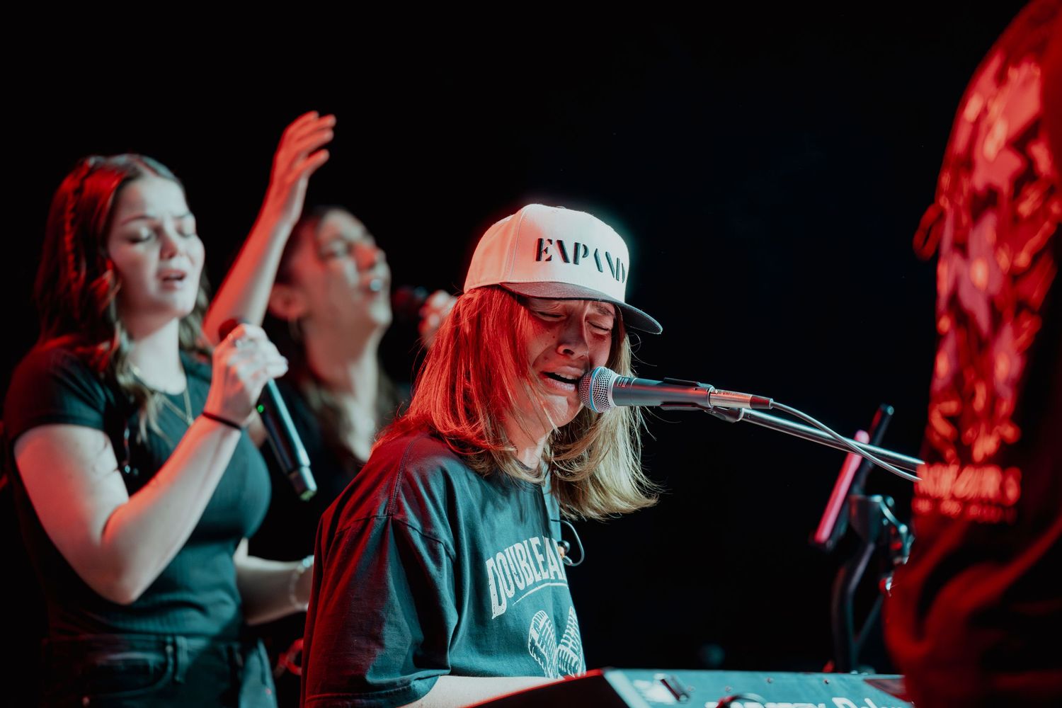 Blonde musician in white cap plays keyboard while backup singers perform on a red-lit stage.