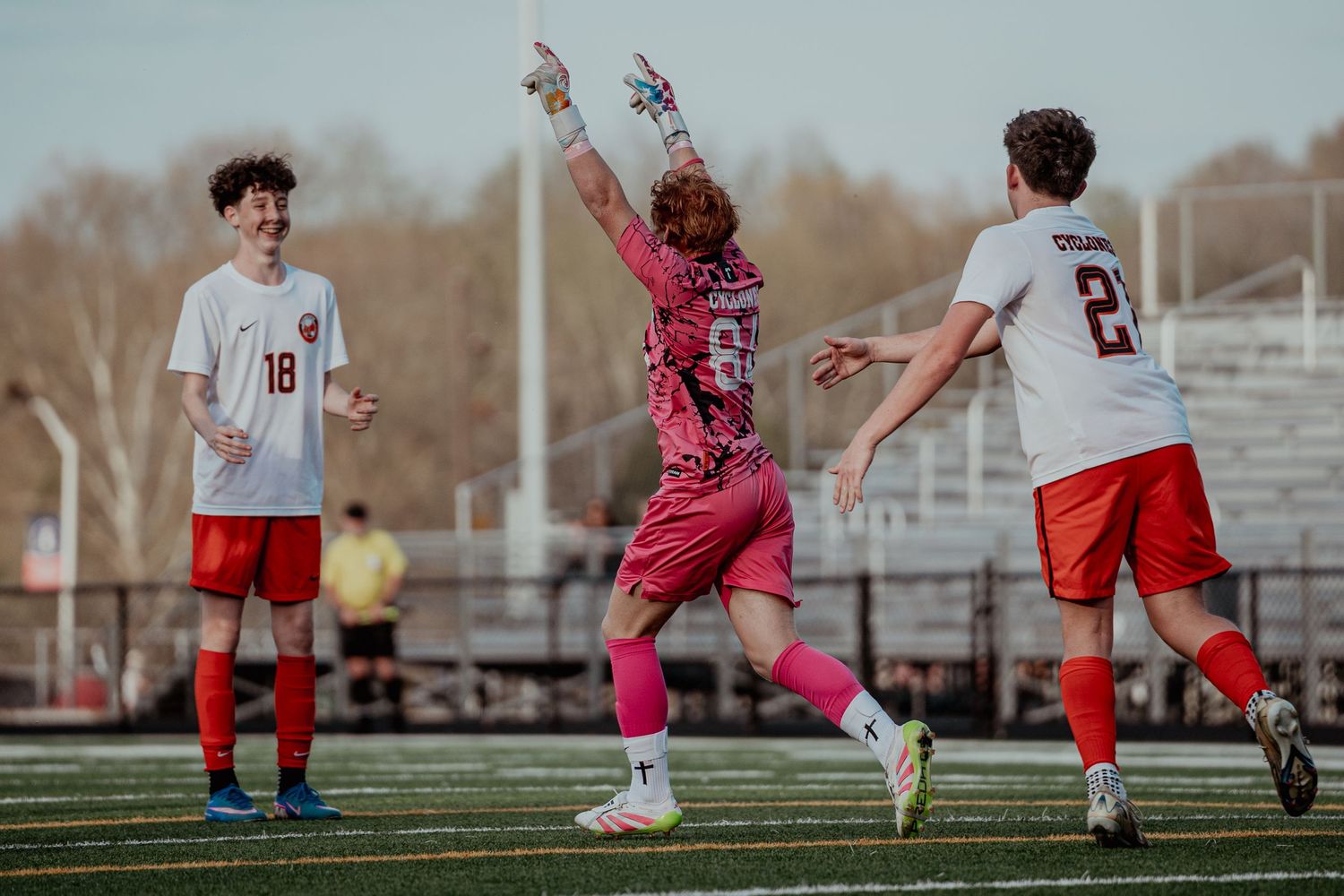 Youth soccer players celebrate on a green field, with a goalkeeper in pink raising arms in victory.