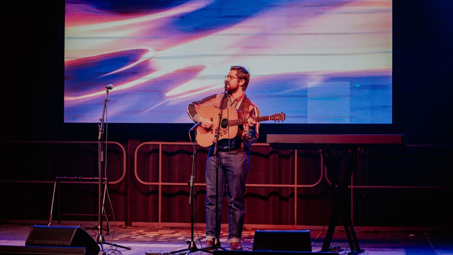 Musician performs on stage with acoustic guitar against colorful illuminated backdrop during live concert performance.