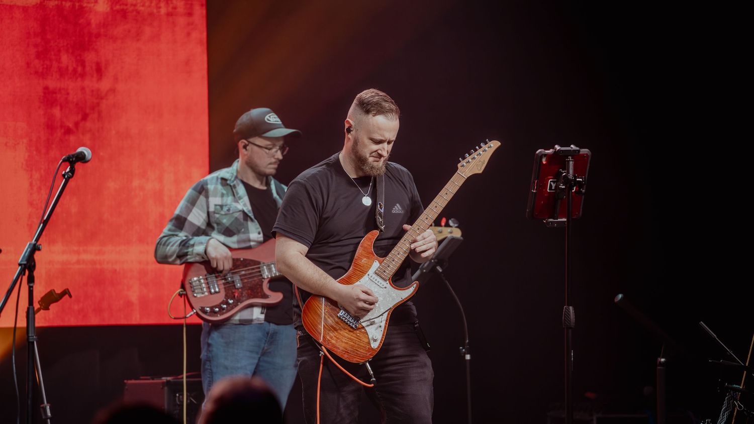 Musicians perform on stage with guitars against a red-lit backdrop during a live concert performance.