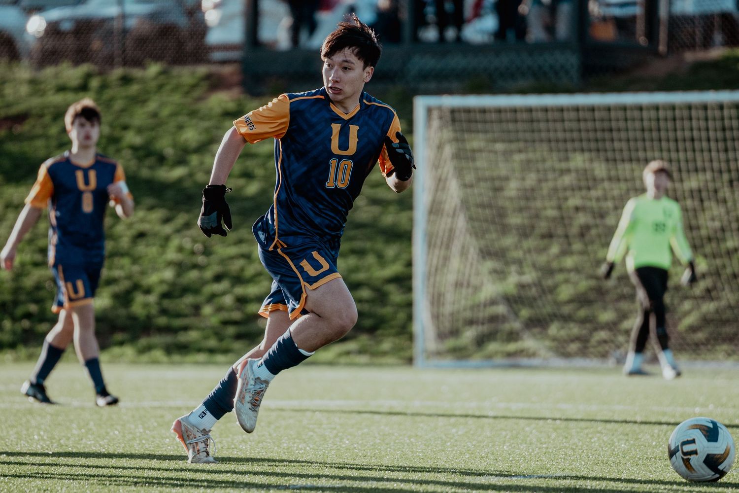 Soccer player wearing number 10 navy and gold jersey dribbles ball on green field during competitive match.