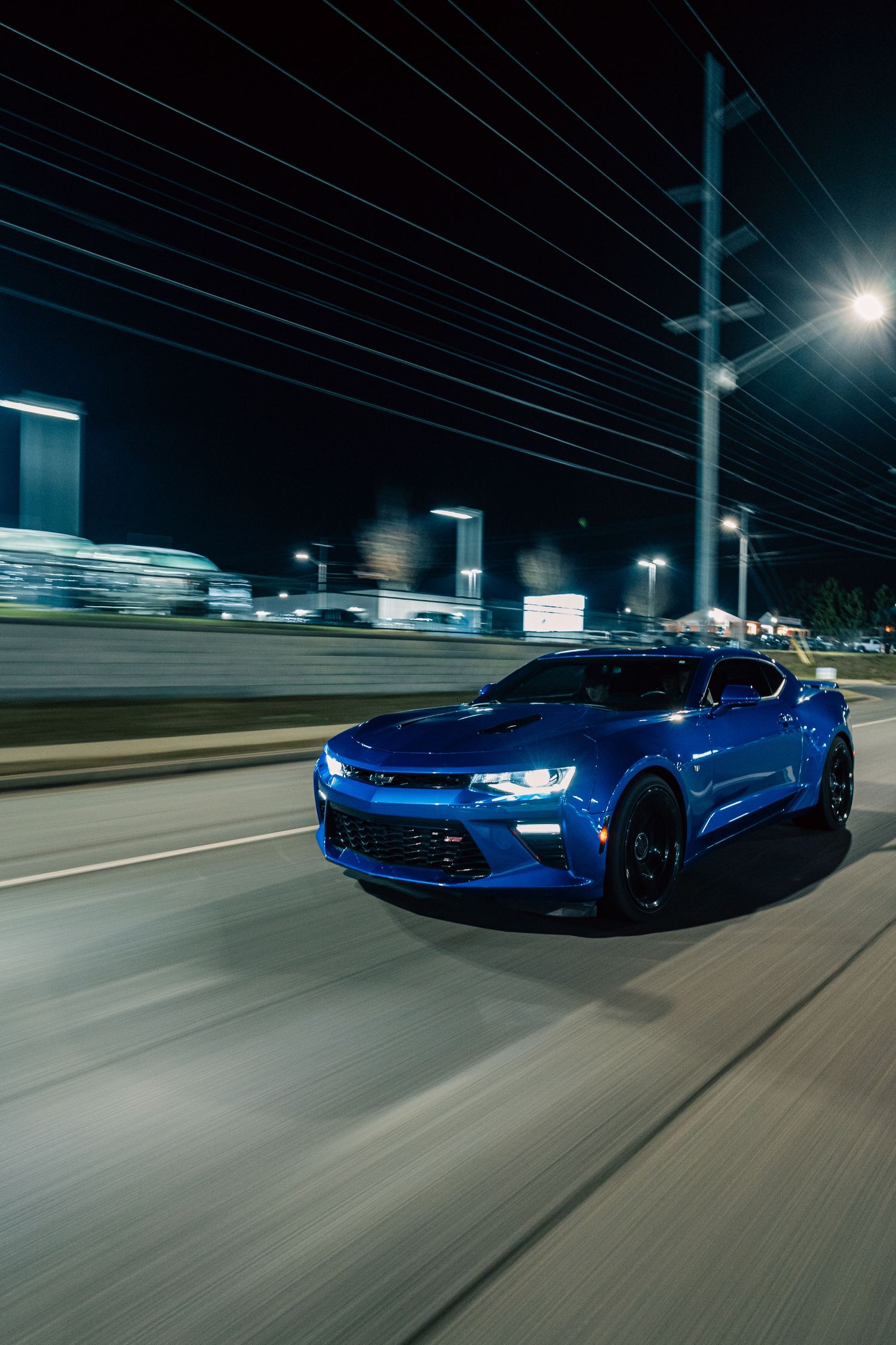 A bright blue Chevrolet Camaro speeds down a highway at night with power lines and city lights in the background.