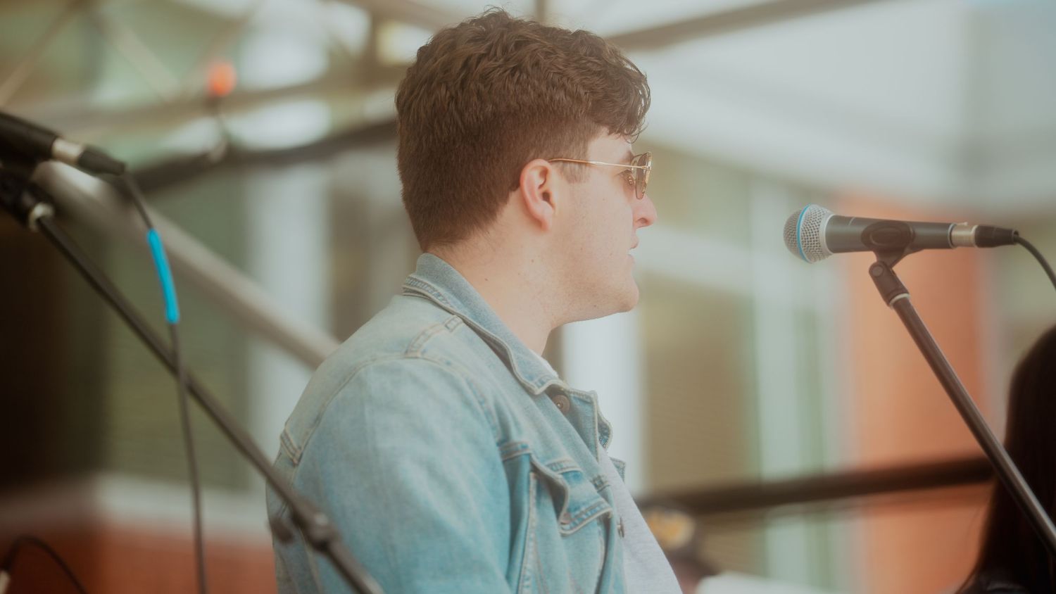A performer in denim shirt speaks into microphone on stage with vintage film-style lighting.