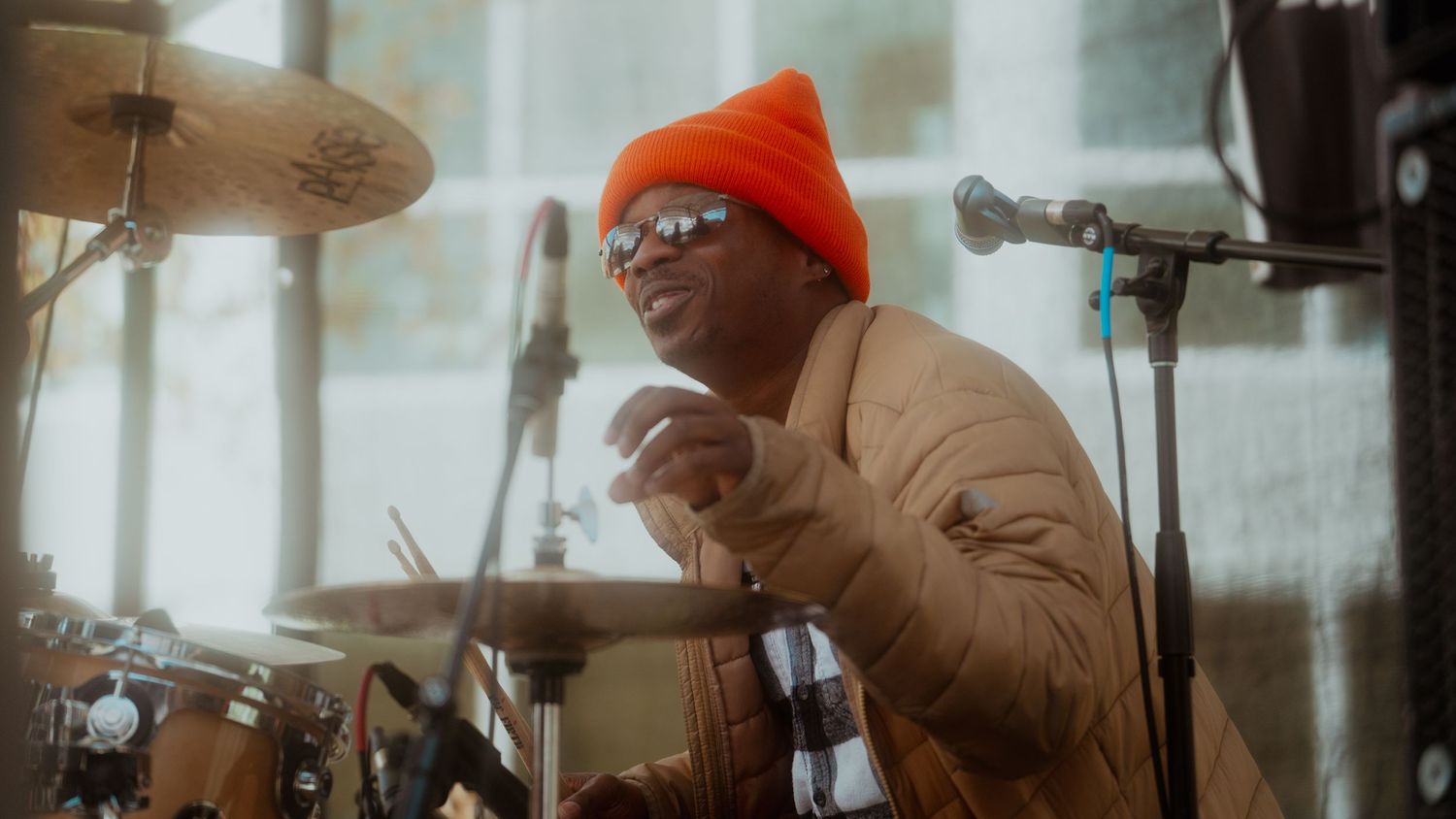 Drummer wearing an orange beanie and glasses performs behind a drum kit in a live concert.