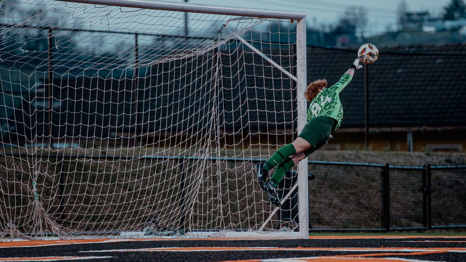 Soccer goalkeeper in green uniform making a diving save near the goal post during a match.