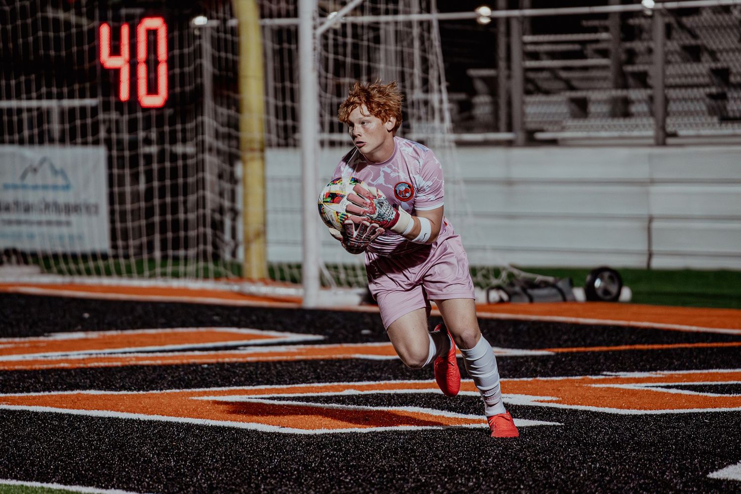 Young redhead goalkeeper in pink jersey catches soccer ball on turf field at night game.
