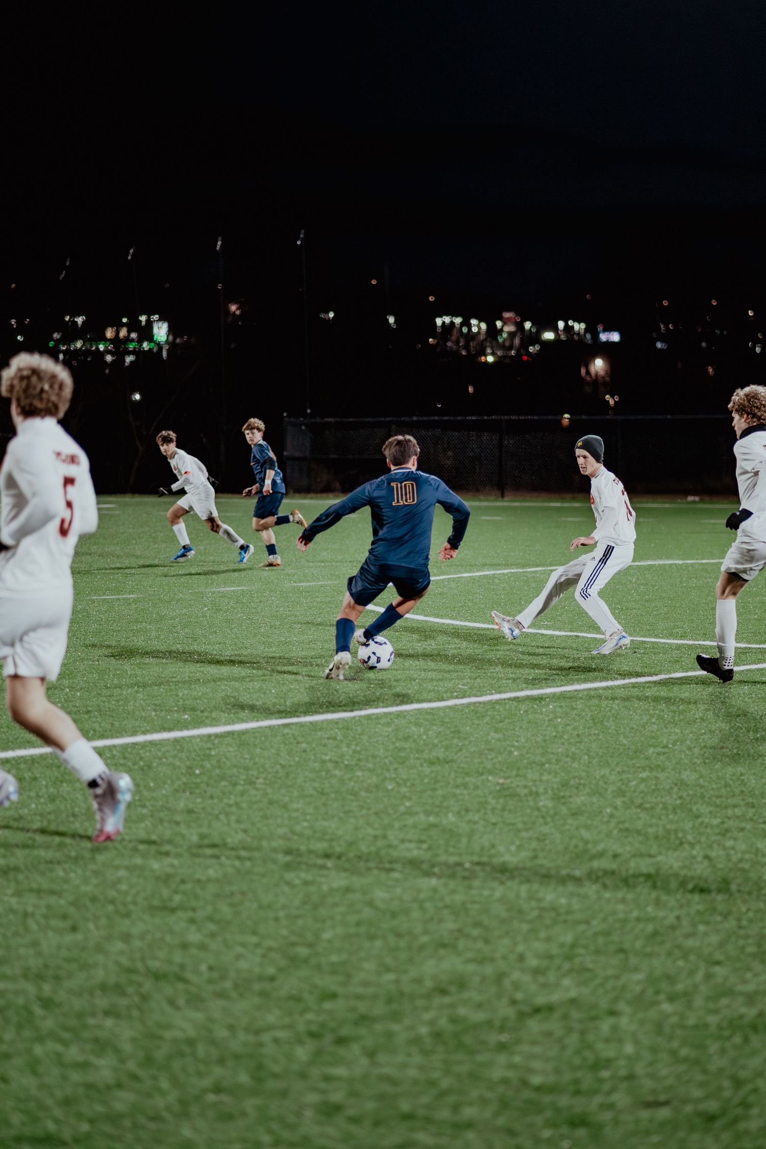 Soccer players compete in a nighttime match on an illuminated artificial turf field under dark skies.