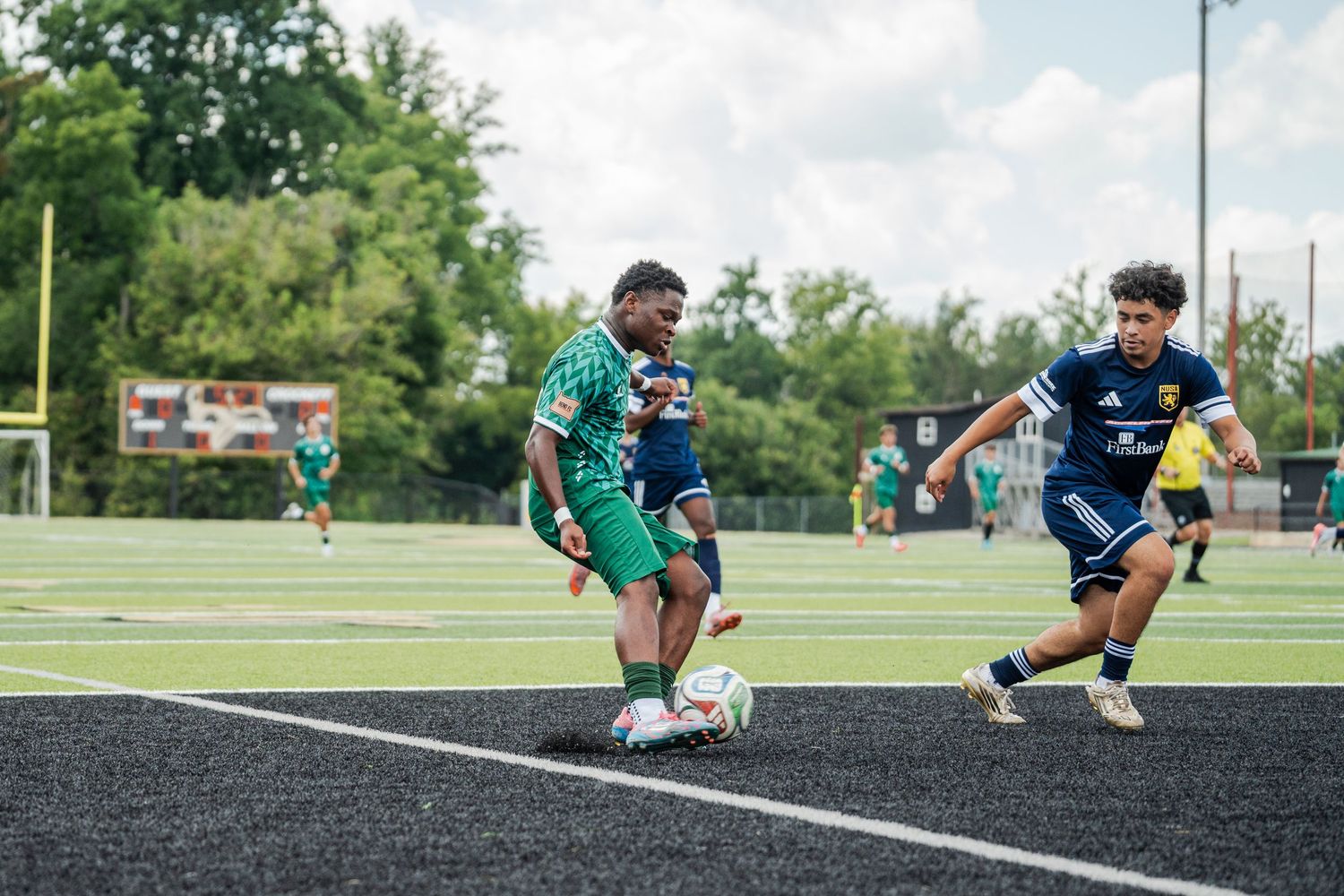 Two soccer players compete for the ball on a turf field during an outdoor match on a sunny day.