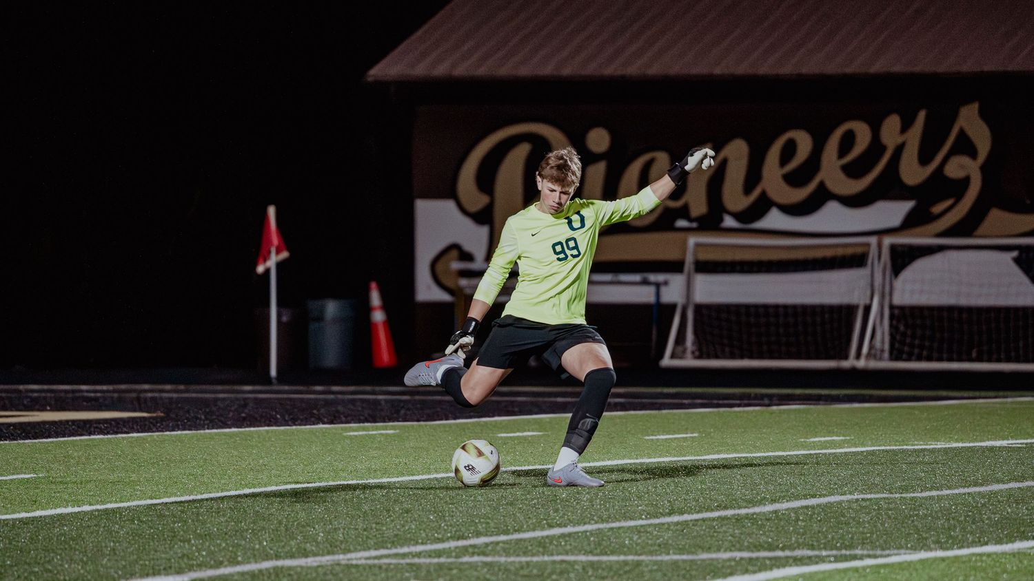 A soccer goalkeeper in yellow uniform prepares to kick the ball on a turf field at night.