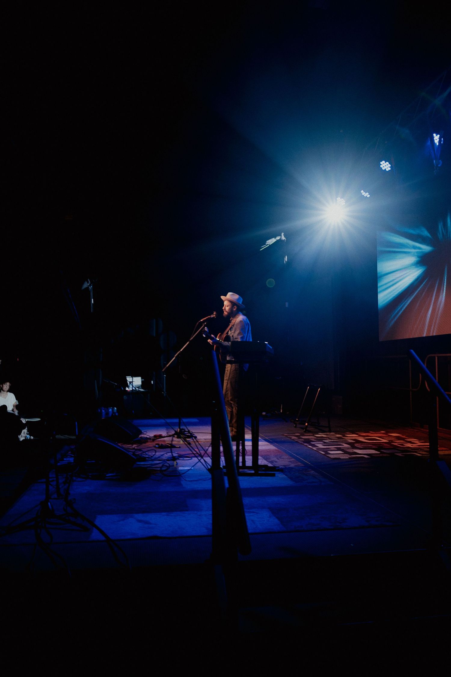 Silhouette of performer standing at microphone on dark stage with dramatic blue spotlight and geometric backdrop.