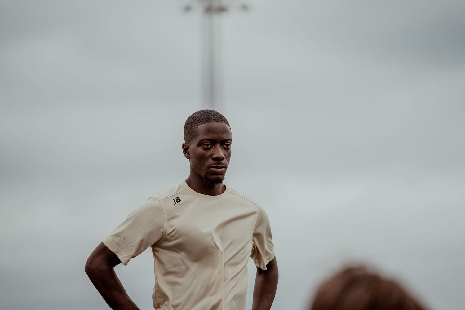 Young Black male athlete in beige t-shirt stands confidently on sports field under overcast sky.