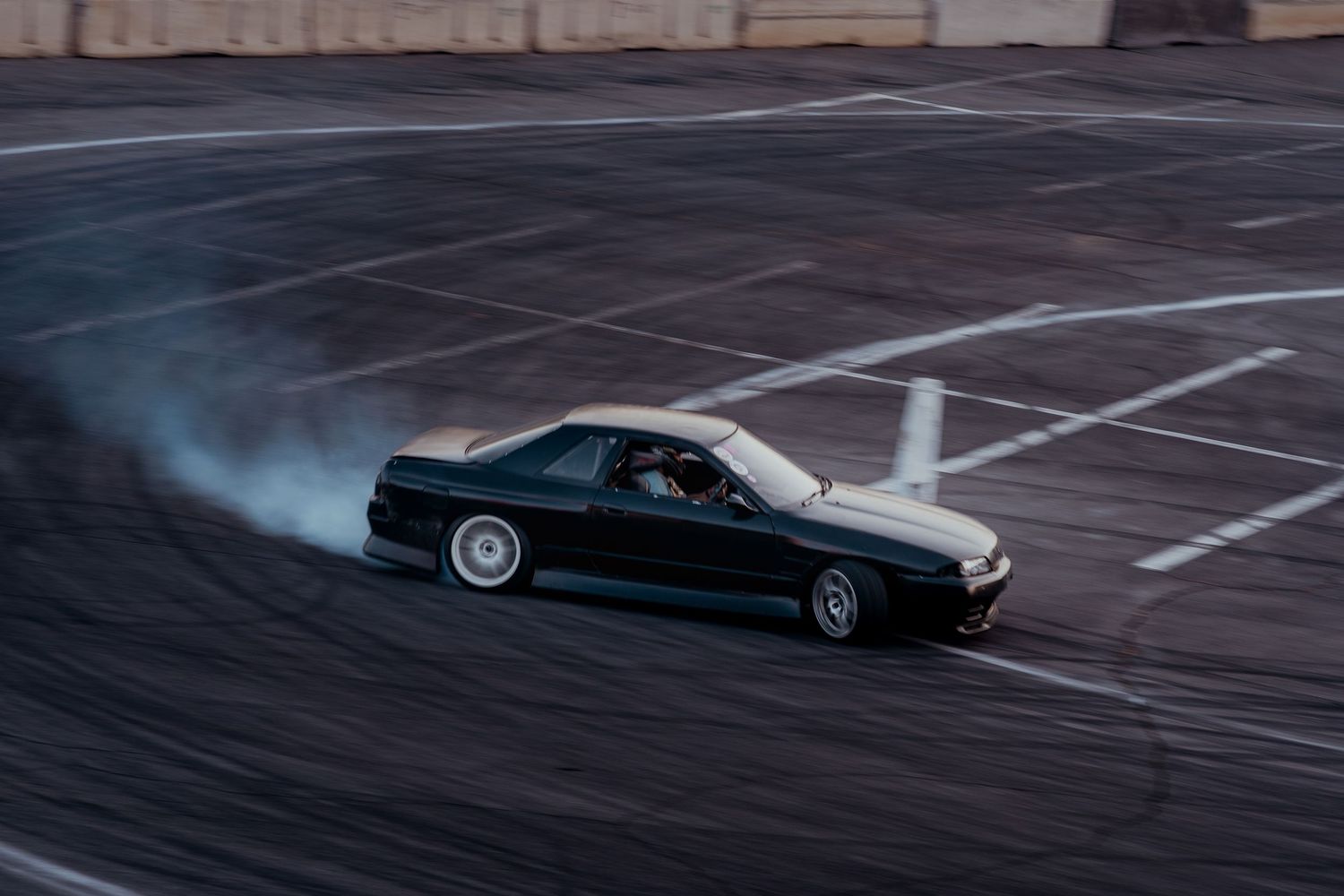 A black sports car drifts on an empty parking lot creating tire smoke during a performance driving demonstration.