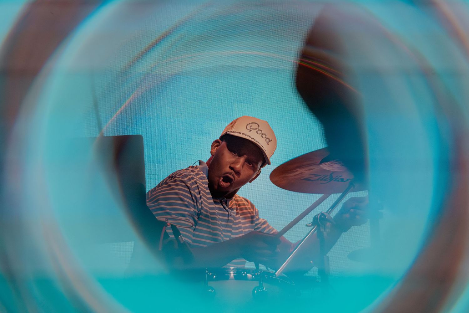 A drummer wearing a striped shirt and cap plays cymbals, photographed through a circular lens for artistic effect.