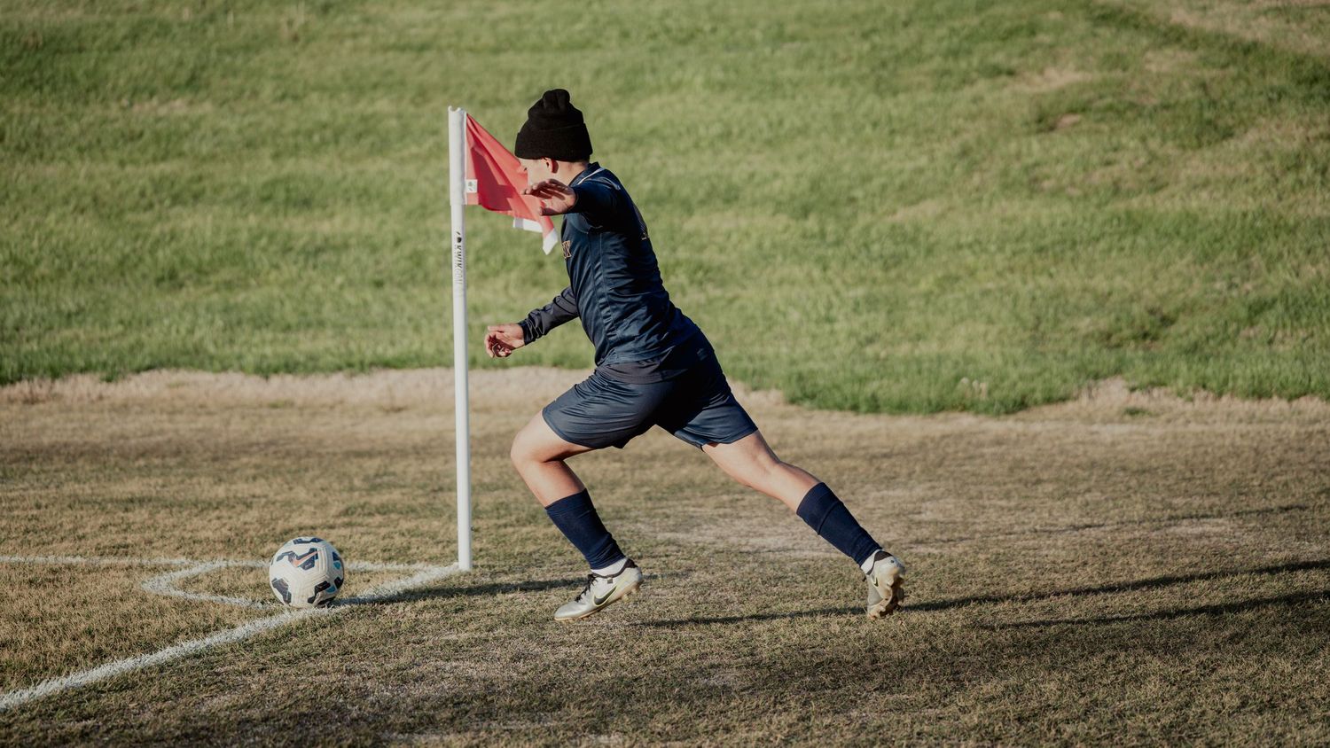 Soccer player in black uniform and knee socks strikes a corner kick on a grass and dirt field next to a flag marker.