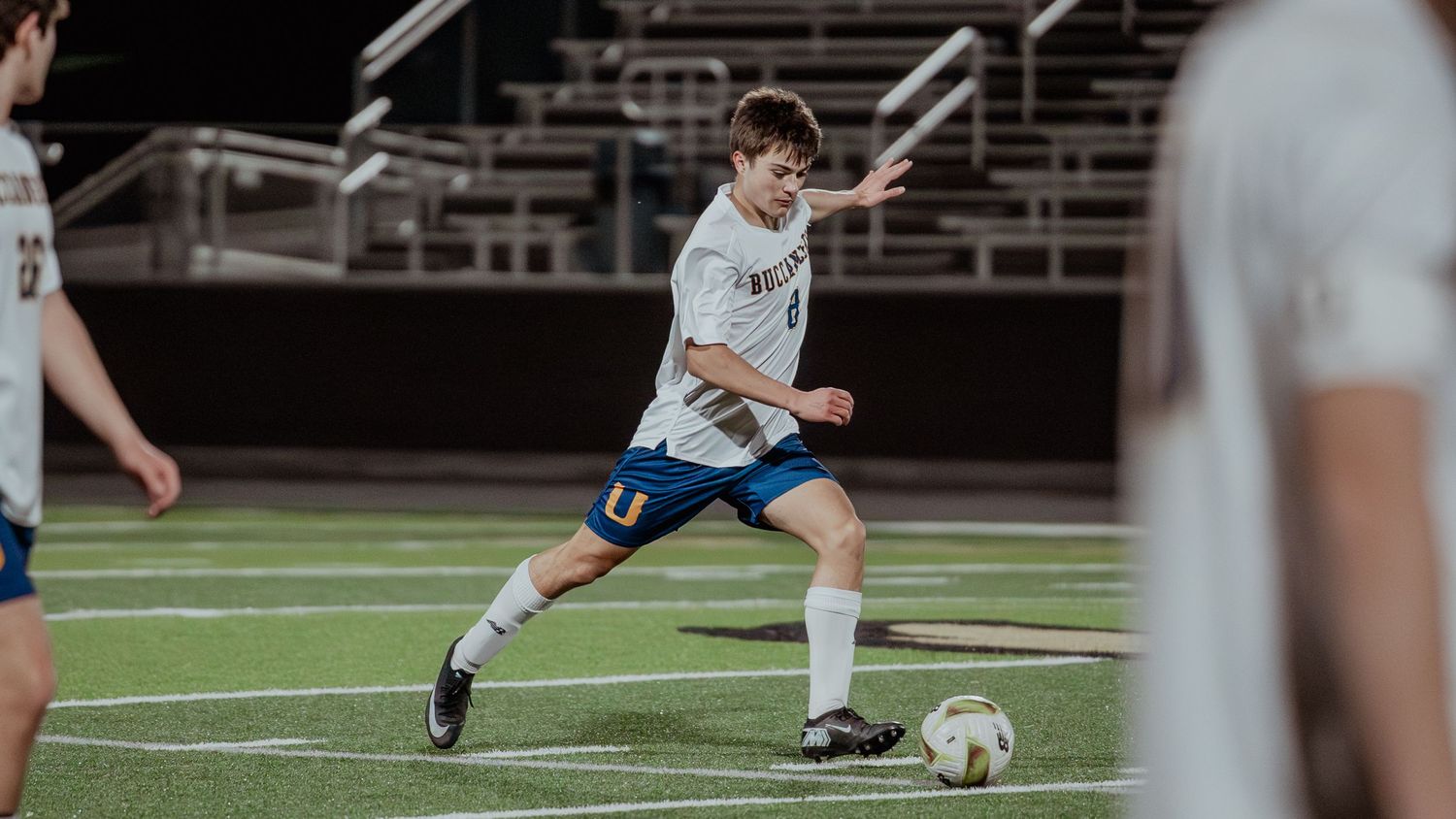 Soccer players in white uniforms compete for the ball on an artificial turf field during a match.