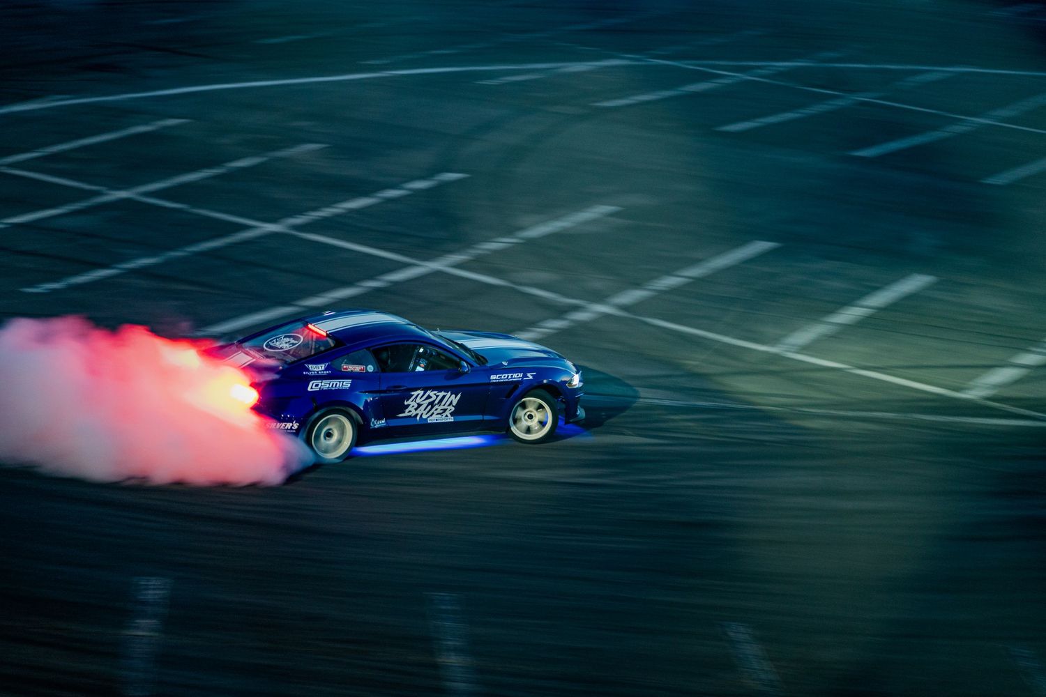 Blue drift racing car performing a tire burn with pink smoke trailing behind it in a dark parking lot.