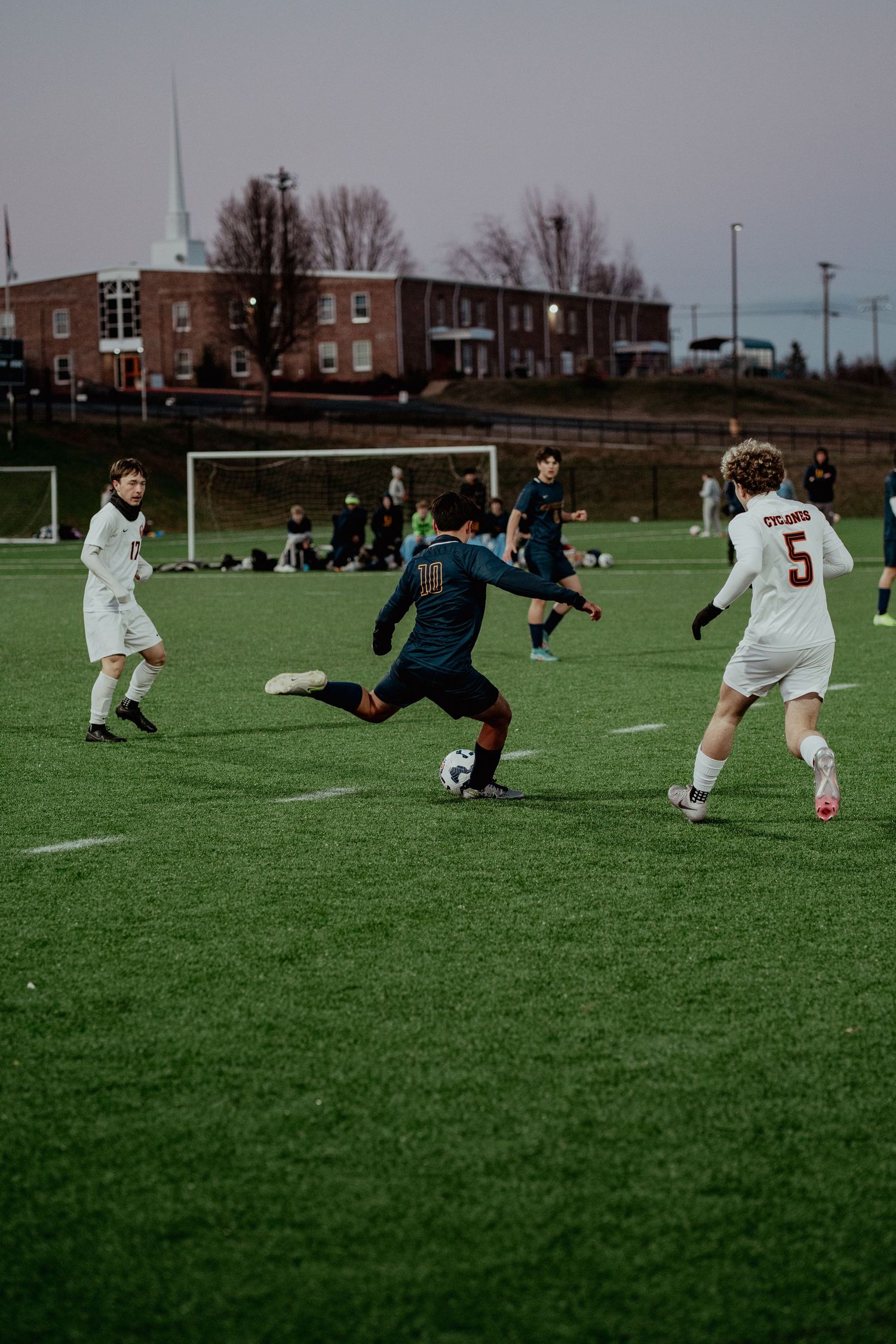 Soccer players compete on artificial turf field with brick buildings visible in background during competitive match.
