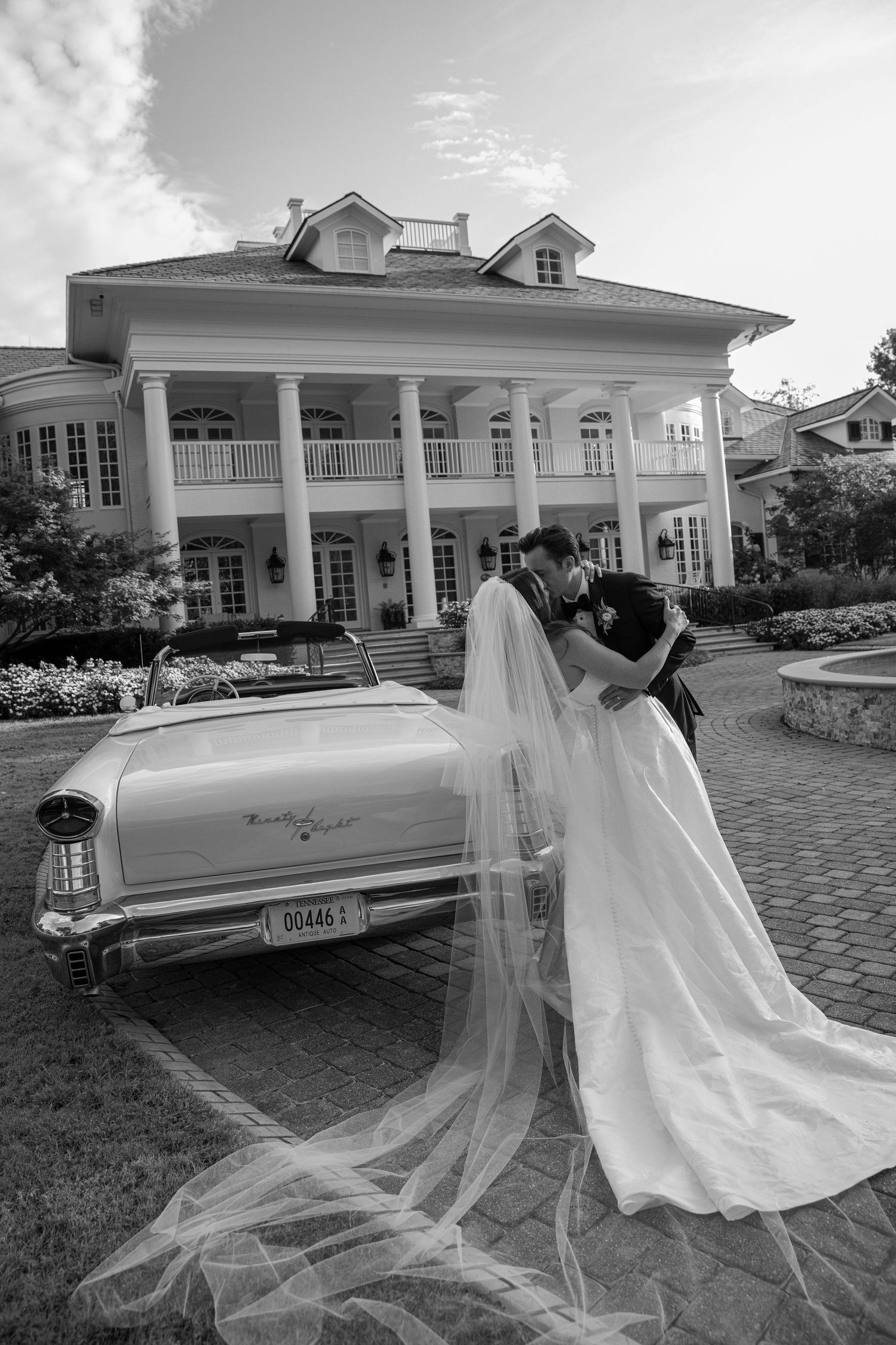 Classic white vintage car parked outside elegant white colonial mansion with long flowing veil trailing behind.
