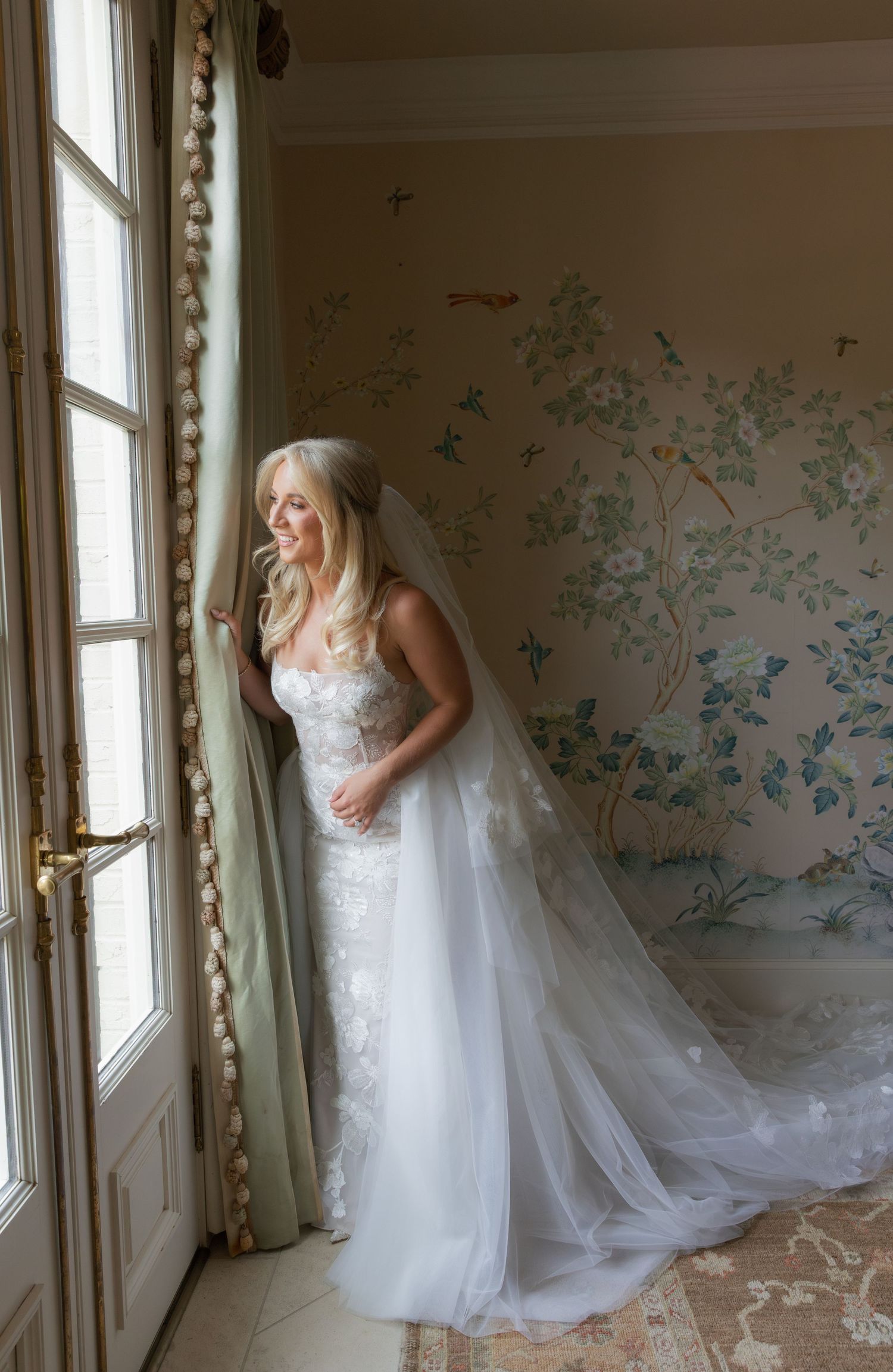 Bride in lace wedding gown and veil stands by ornate French doors with floral wallpaper backdrop.