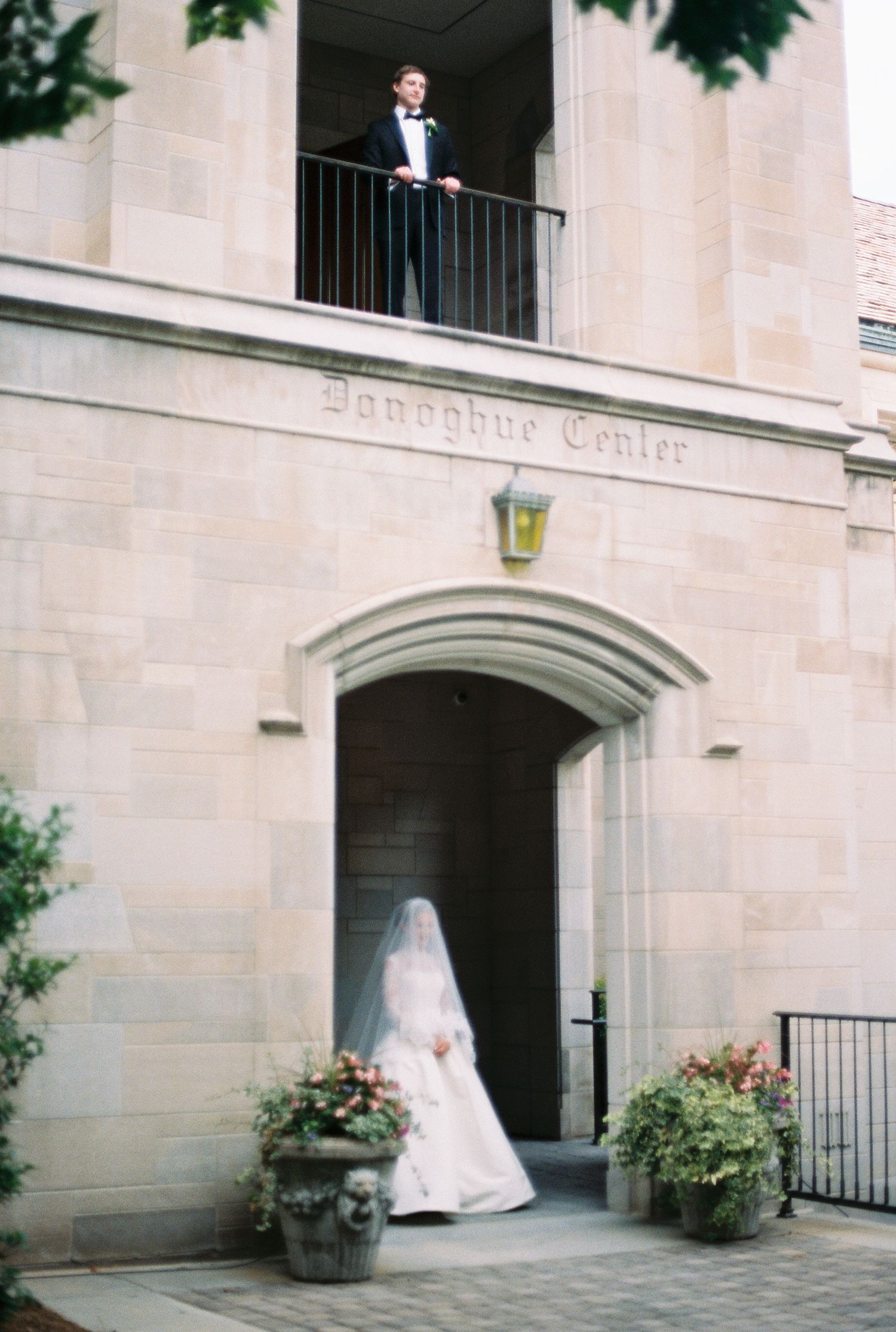 Wedding moment captured at a stone building entrance with a bride below and groom looking down from a balcony.