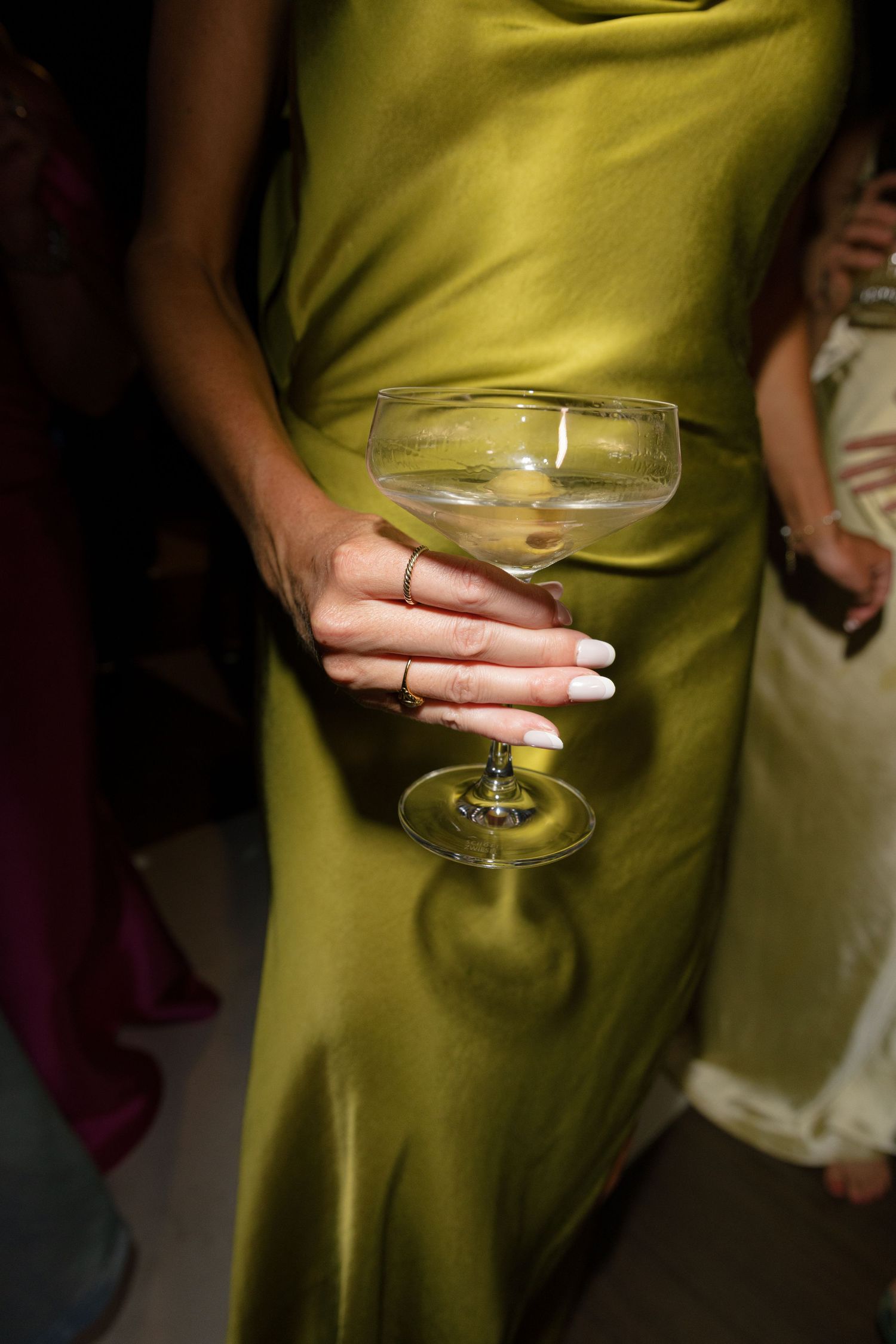 A guest in an olive green satin dress holds a champagne coupe glass at a wedding party.